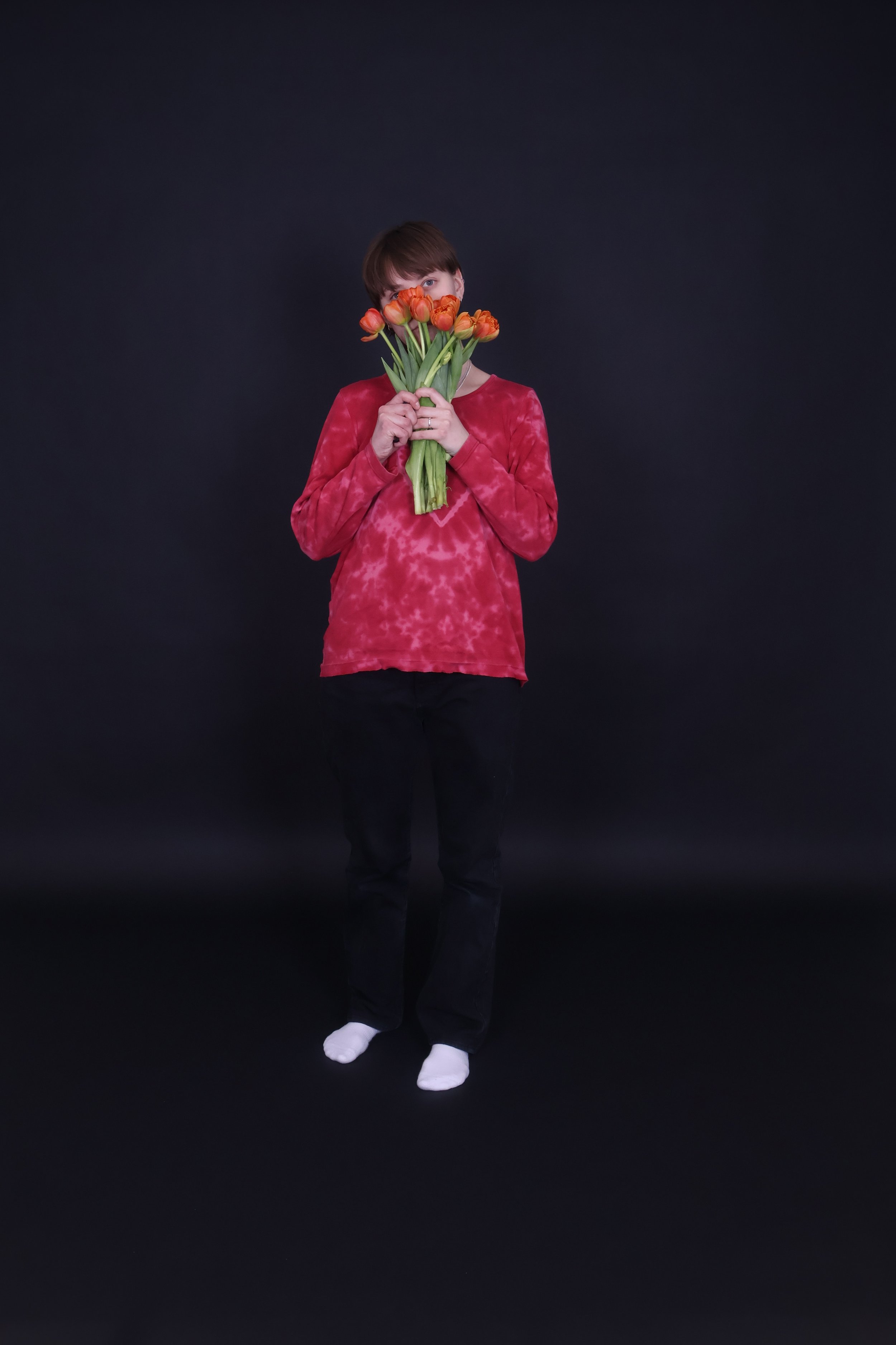 Nuoren henkilö seisoo mustalla taustalla, pitelee kimpua oransseja tulppaanin kukkia kasvonsa edessä.