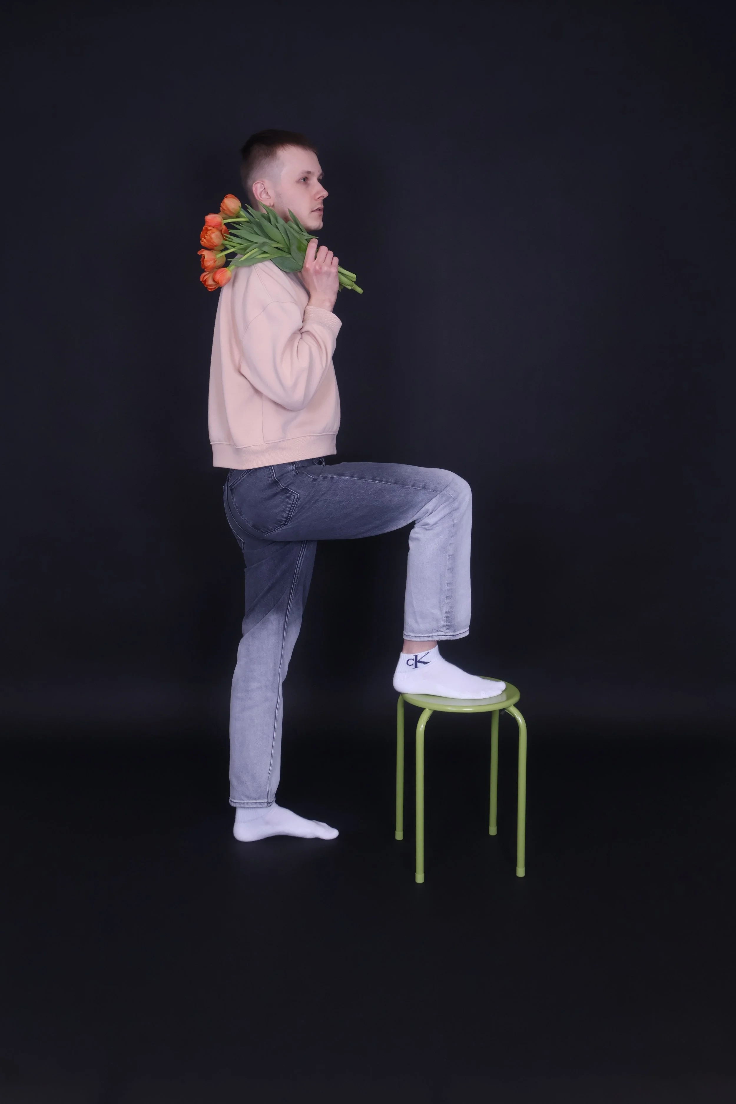 Mies seisoo yhdellä jalalla tuolilla ja pitää kukkakimppua olkapäänsä takana.