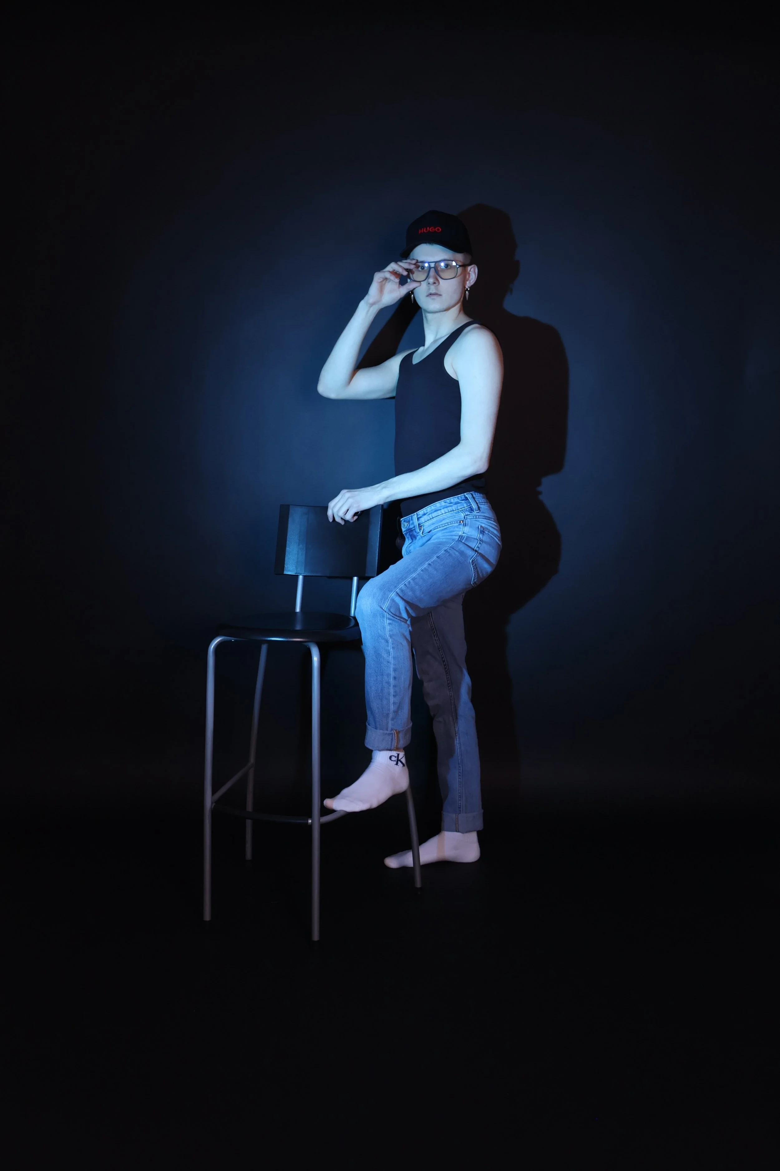 Nuori mies seisoo tuolilla pimeässä taustassa, hänellä on musta tanktop, farkut, lippis ja silmälasit.