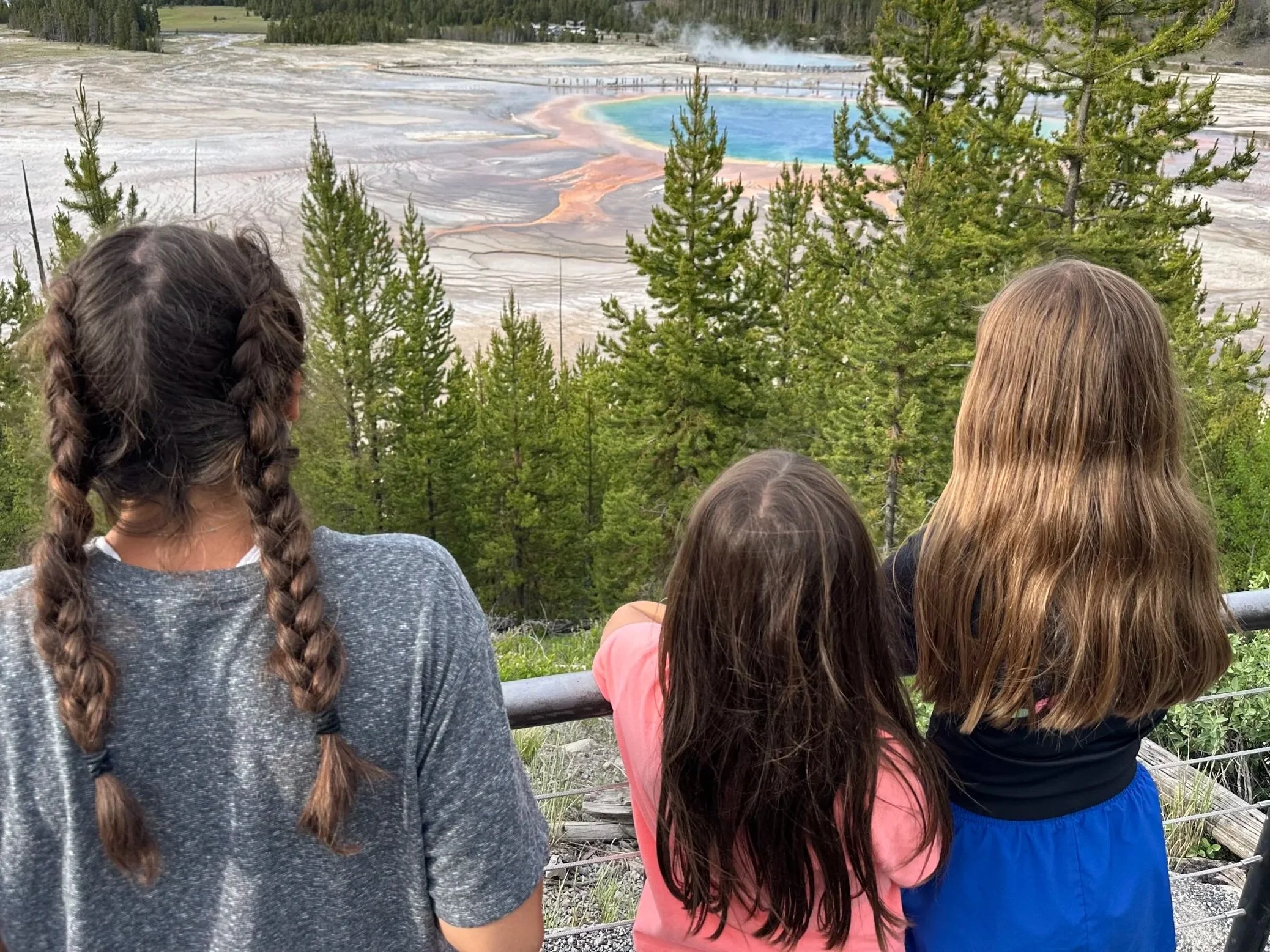Three children with braided and long hair looking at a colorful geothermal pool surrounded by trees and steam rising from it.
