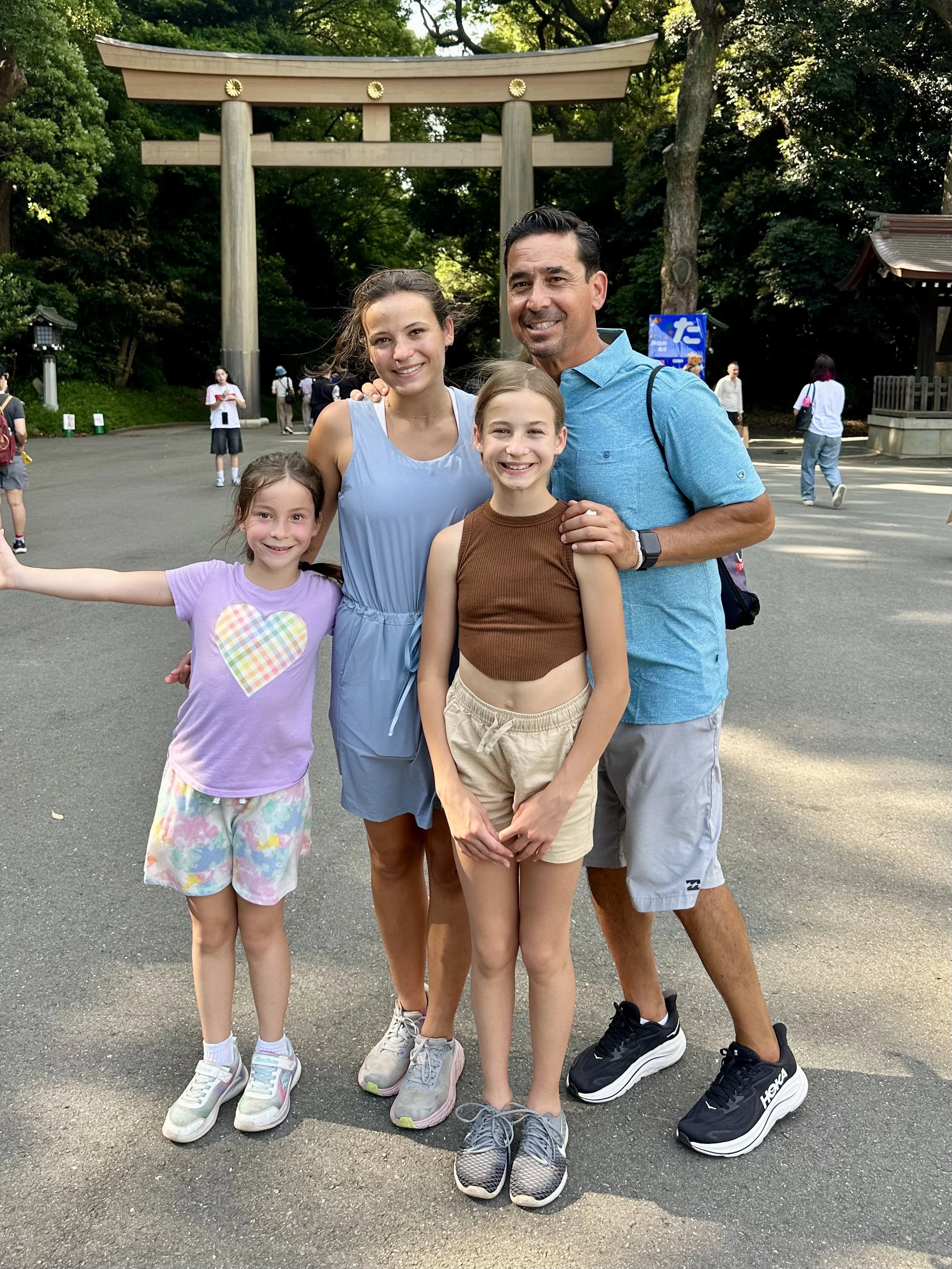A family with three young girls and their father standing in front of a traditional Japanese torii gate at a park or shrine, surrounded by trees.