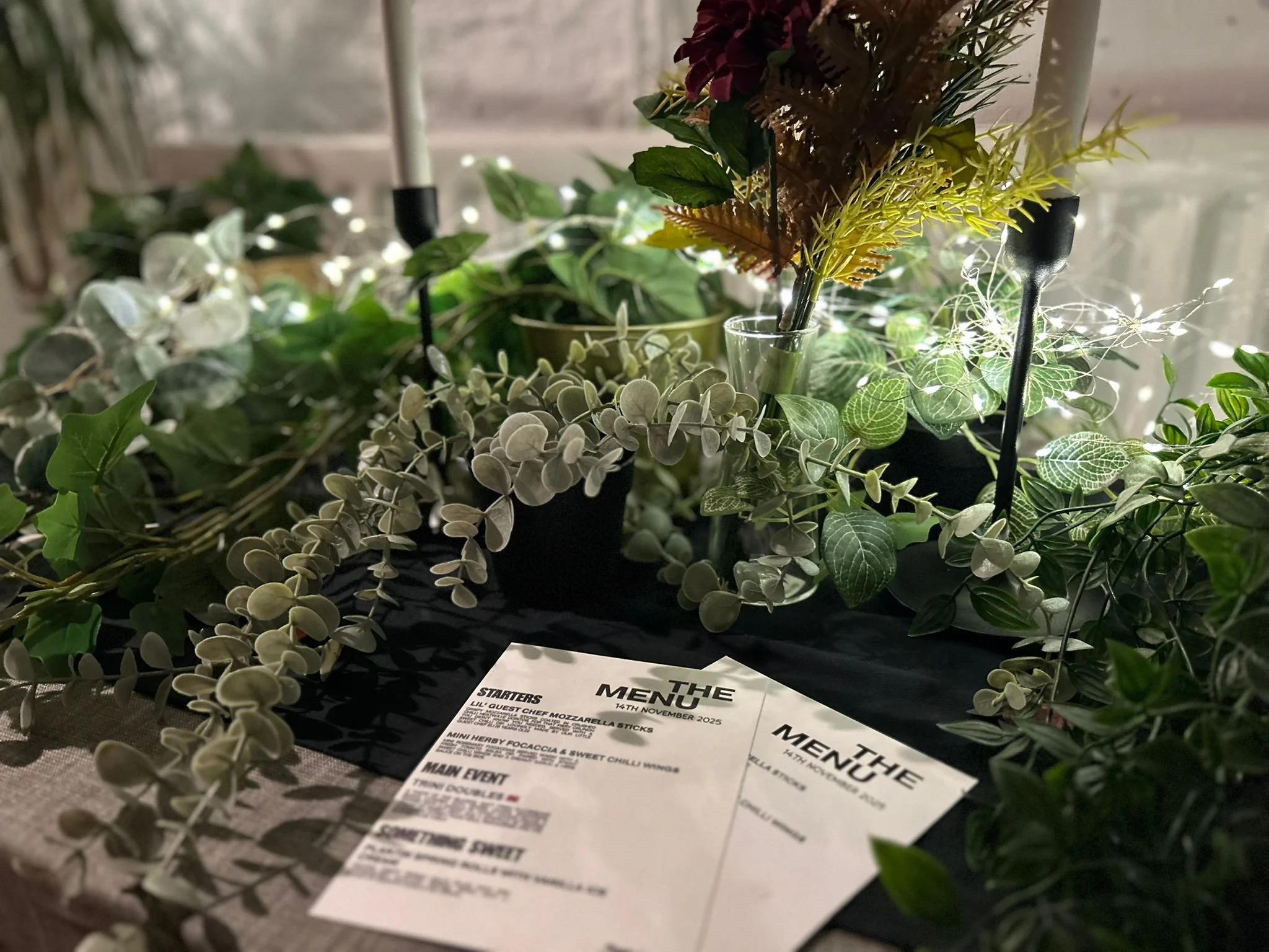 A table with a black table runner decorated with green and white artificial plants and foliage, two candles, and two printed menus from OK DENI CIC's supper club event
