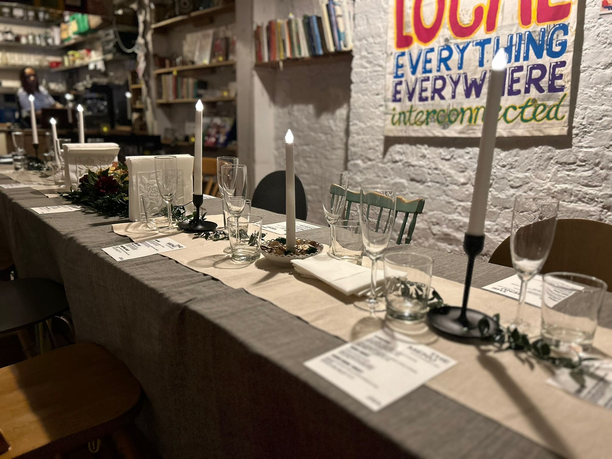 A decorated dining table with candles, glassware, and place cards, set in a cozy indoor space with bookshelves and a colourful quilt on the wall. The Hornbeam CIC