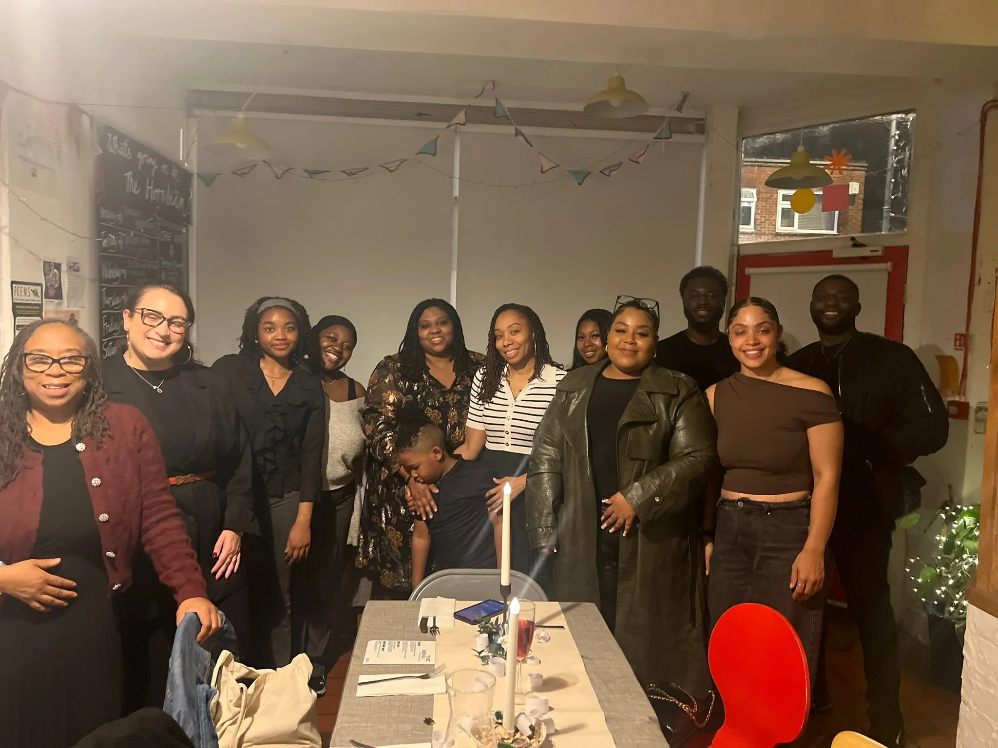 Group of twelve people gathered in a room, posing for the photo with smiling faces, some standing behind a table with a candle, papers, and drinks, decorated with hanging paper flags and balloons.