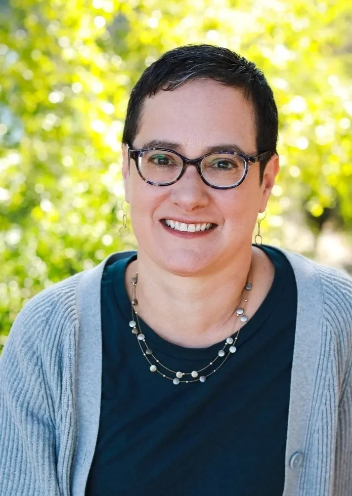 A woman with short dark hair, glasses, and earrings, smiling outdoors with a background of blurred green foliage.