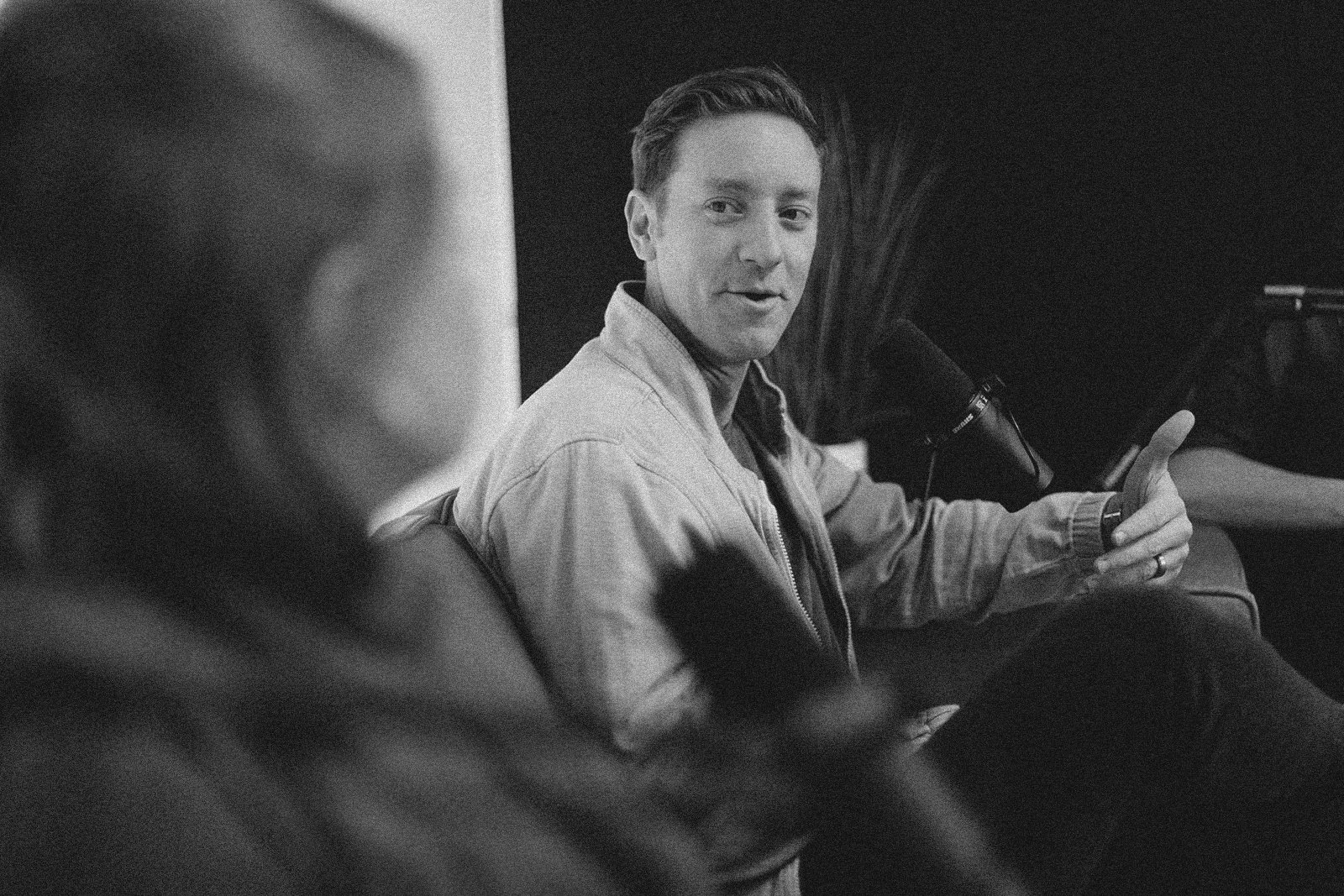 A man with short hair, wearing a jacket, is seated and talking to a blurred woman in the foreground. He is giving a thumbs-up gesture. They are in a room with dark walls and a plant in the background.