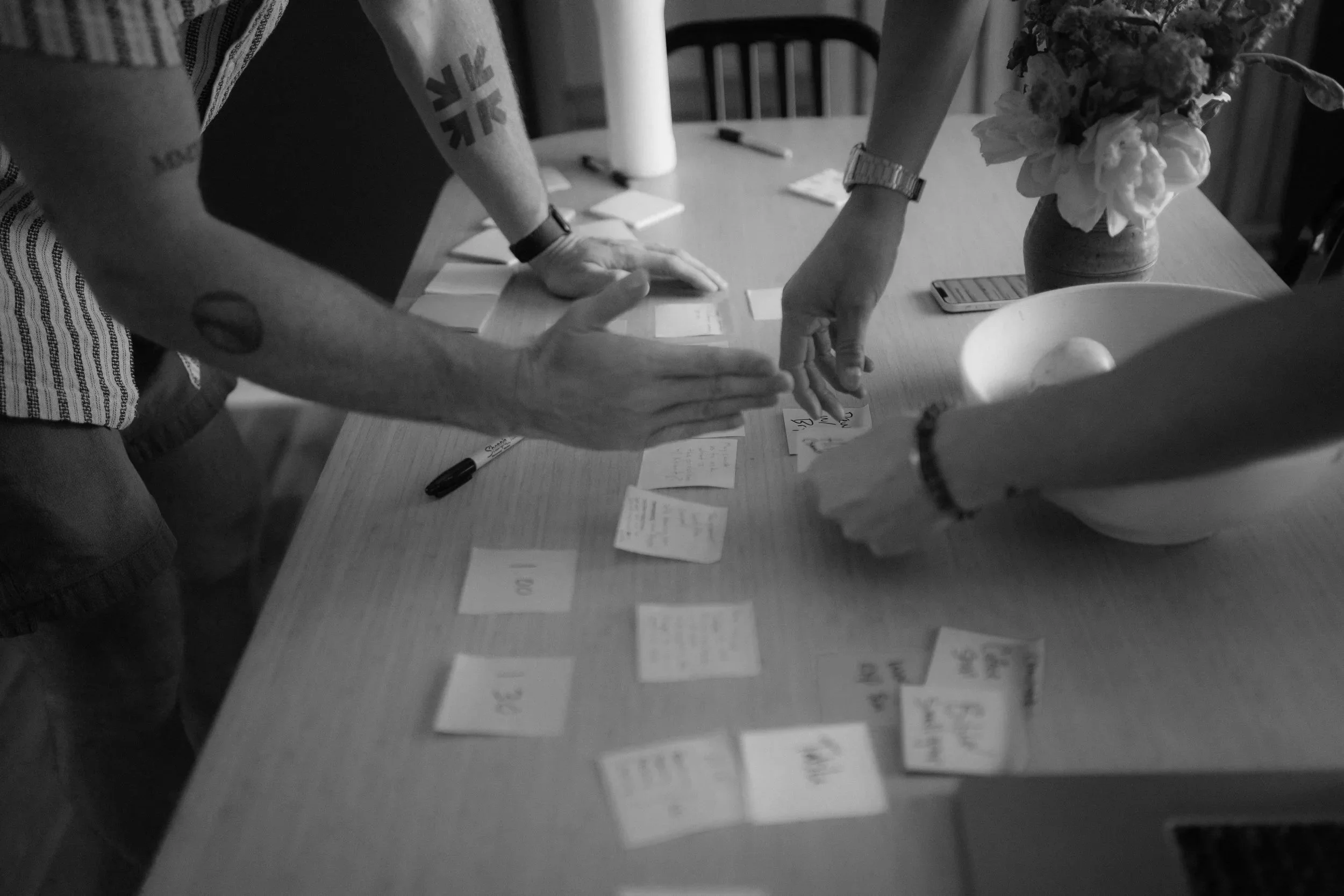 People gathered around a table with scattered notes, a pen, a smartphone, a cup, a vase with flowers, and some props, with one person reaching for notes and another placing cards on the table.