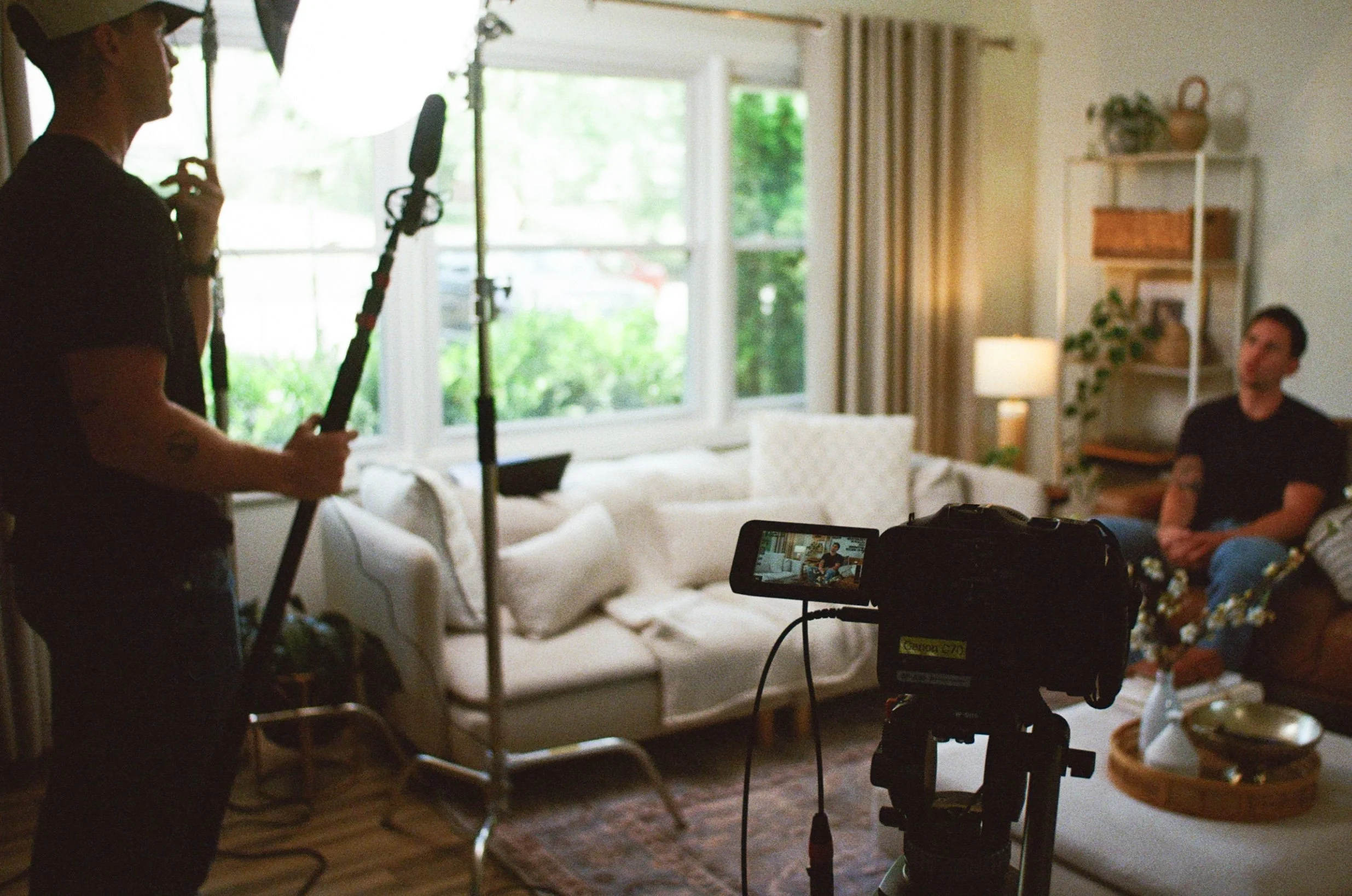 A person holds a boom microphone in a well-lit living room with large windows, a white couch, and a bookshelf. Two other people are sitting on the couch, and filming equipment, including a camera and monitor, are set up in the room.