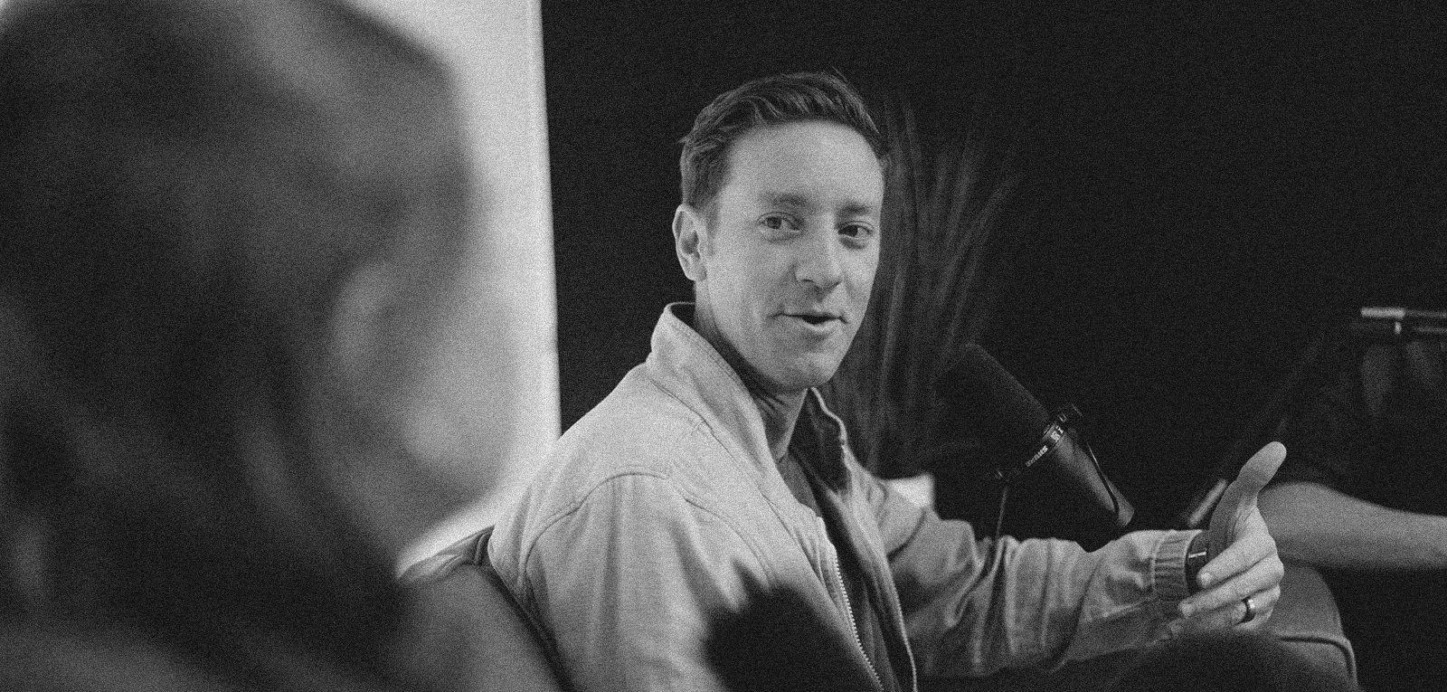 A man with short hair talking into a microphone during a conversation, with a woman in the foreground out of focus in a black and white photo.