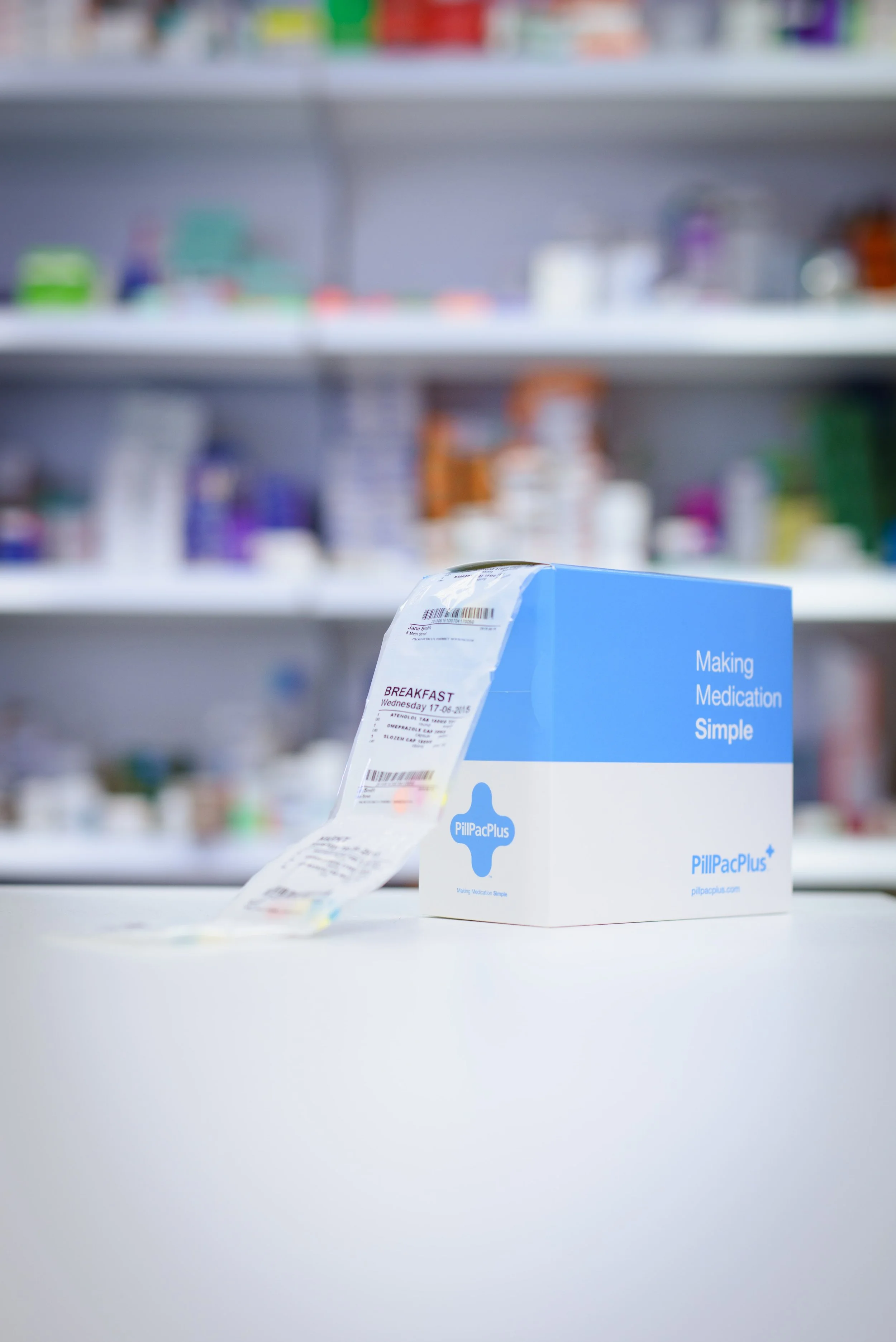 Blue box labeled PillPacPlus with a prescription label attached, placed on a white surface with shelves of medication bottles blurred in the background.