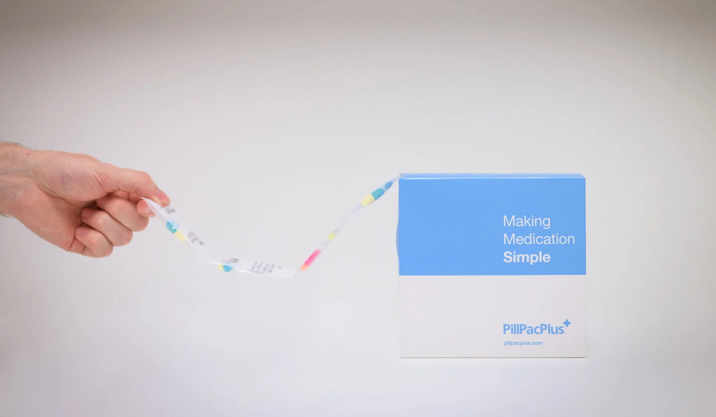 Person holding a strip of colorful pills in front of a white box labeled "Making Medication Simple" by PillPacPlus.