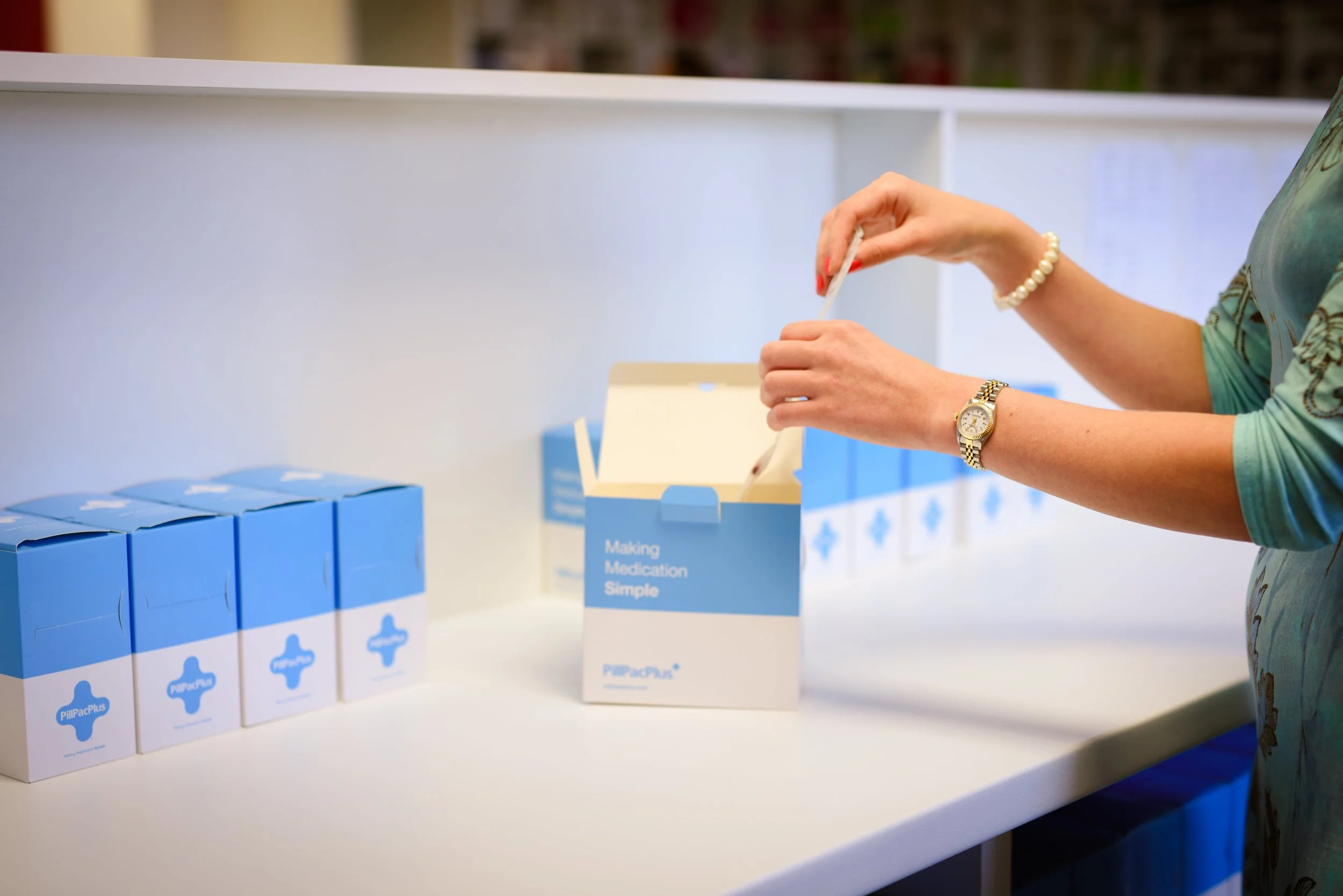 Person with a pearl bracelet and gold watch is handling pills in a white box at a table, with blue boxed medications in front and a white box with blue text reading 'Making Medication Simple' behind.