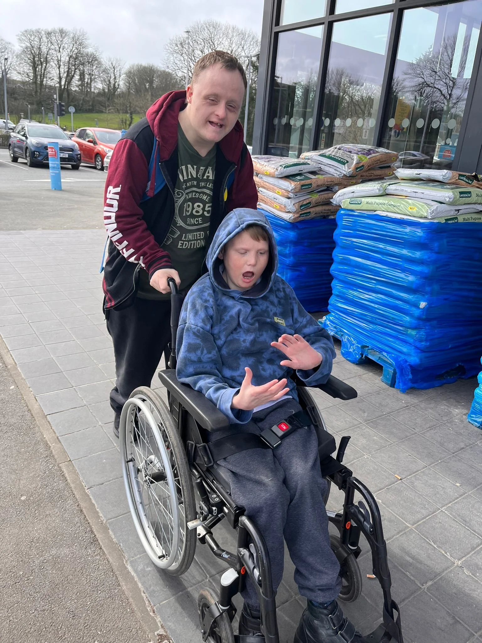 A man pushing a child in a wheelchair outside a store with stacks of compost bags in the background.