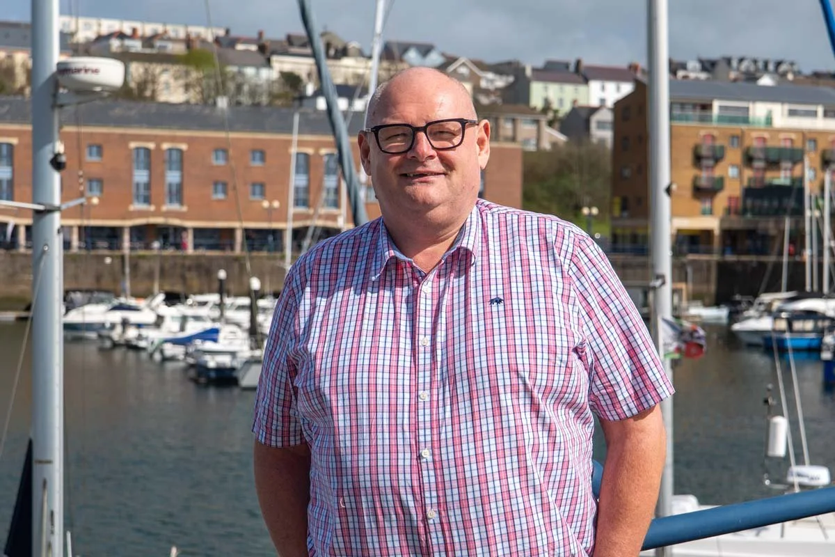 Tom Hale, Director of Silver Springs Support, smiling at the camera, standing near boats docked at a marina with houses in the background.