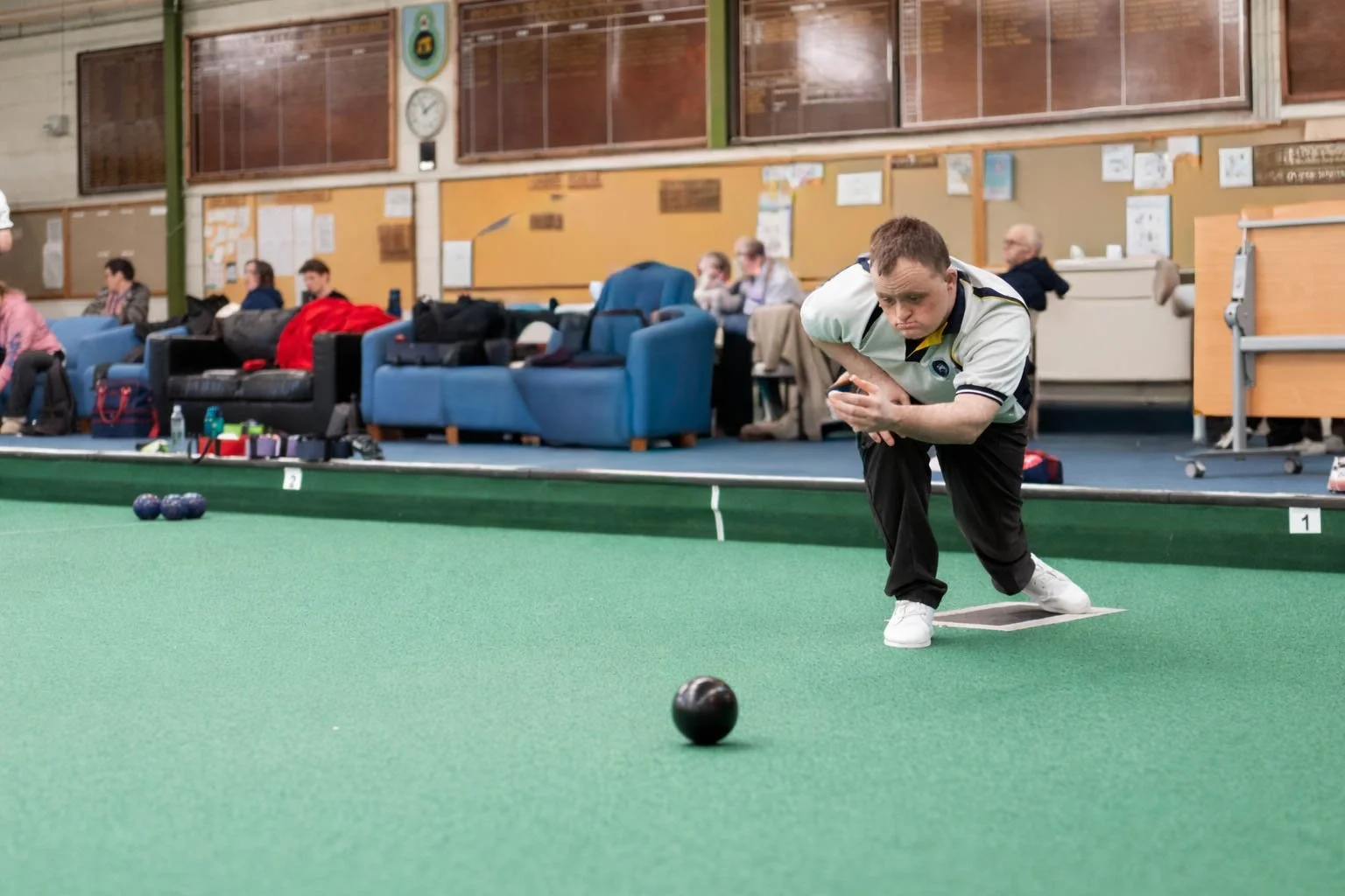 A man playing indoor bowling, with several people seated in the background.