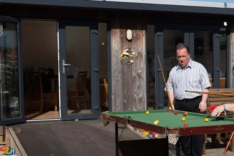 A man playing pool on a green billiards table outside a house during daytime.