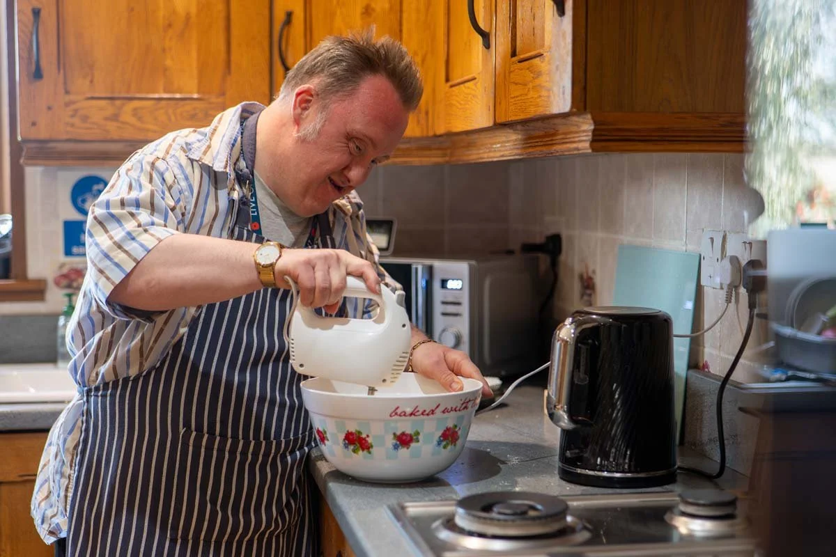 A man wearing a striped apron and a watch, smiling while using a hand mixer in a bowl in a kitchen.