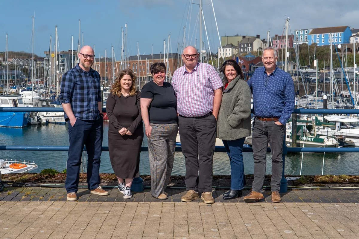 Six people standing in front of a marina with boats and colorful buildings in the background.