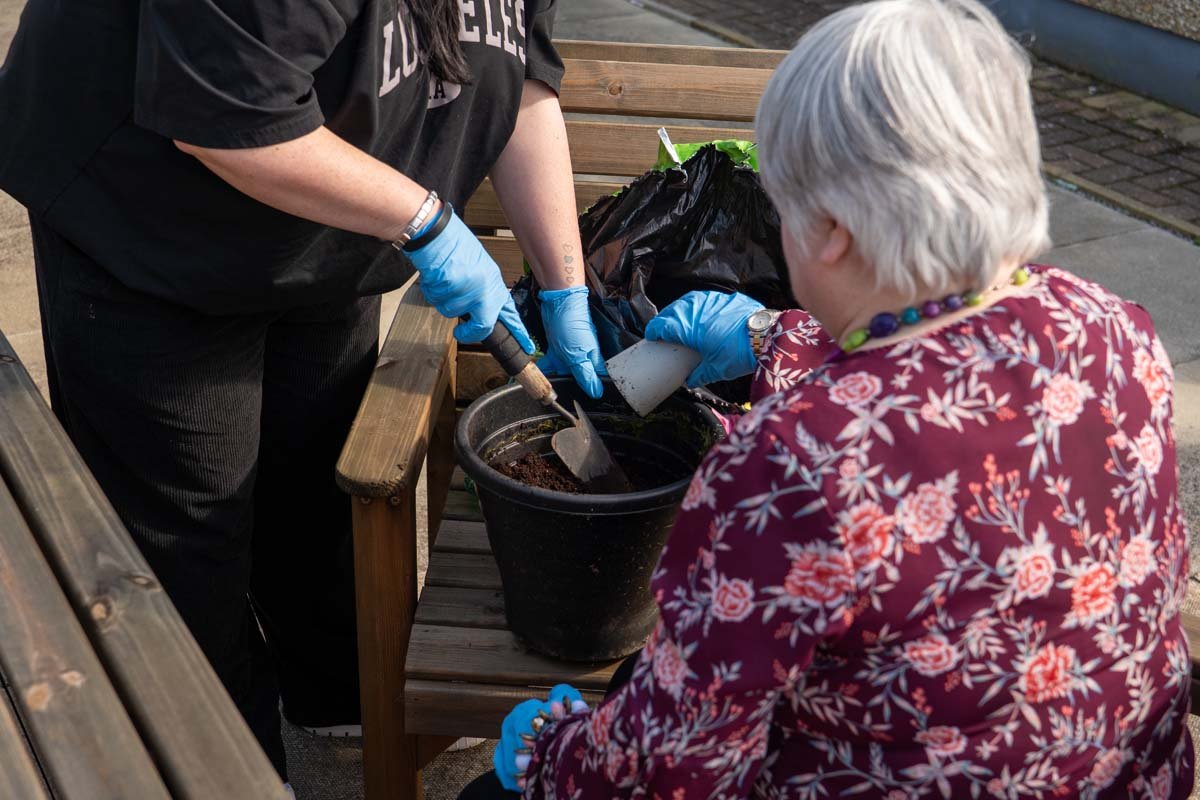 Two women planting flowers in a black pot outdoors, one wearing a black shirt and gloves, the other an embroidered red floral blouse and a colorful beaded necklace, with gardening tools and a black bag on the bench.