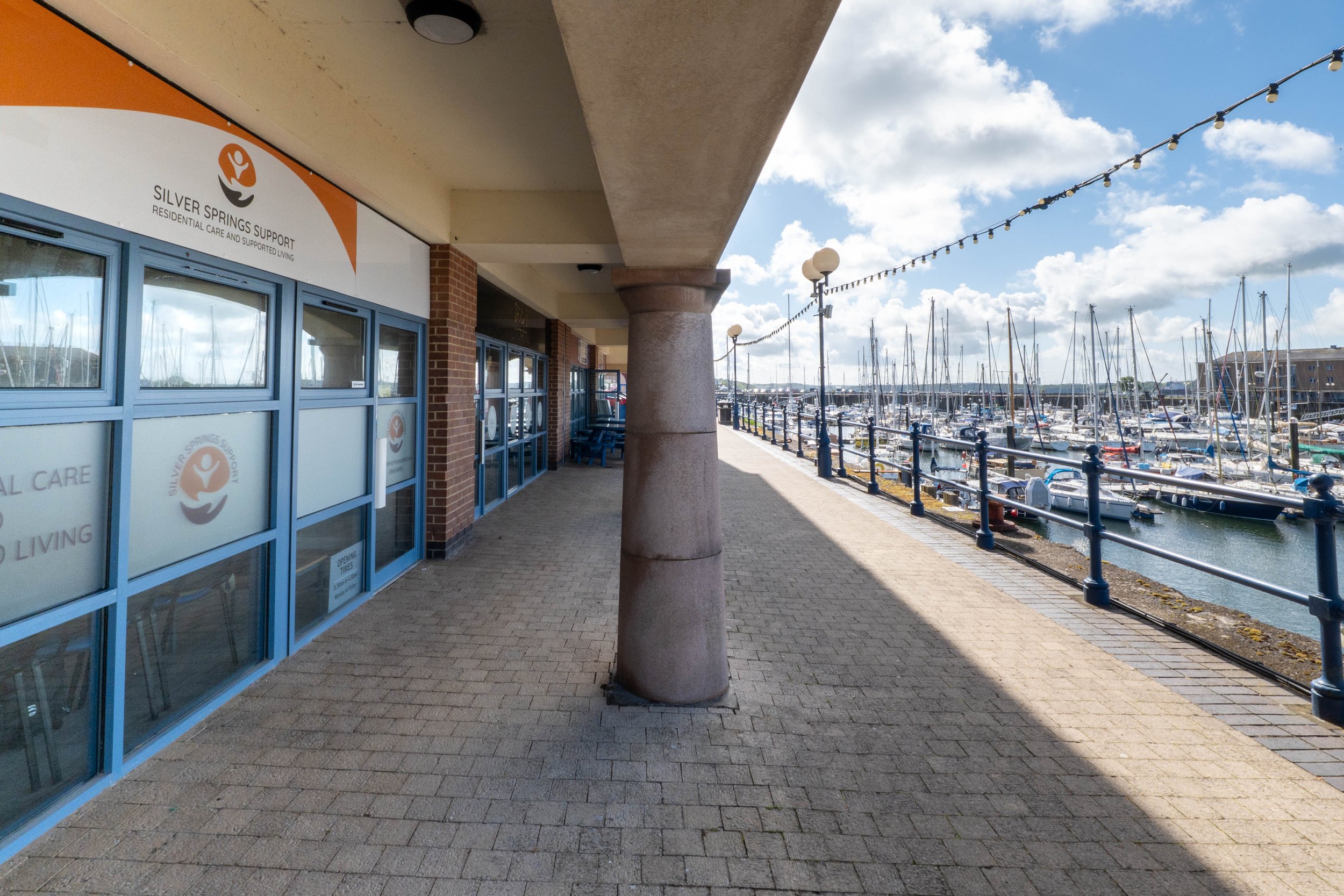 Storefront with glass windows and a sign that reads 'Silver Springs Support Residential Care and Supported Living,' located beside a marina with sailboats and yachts docked along the water. The sky is partly cloudy with string lights hanging overhead.