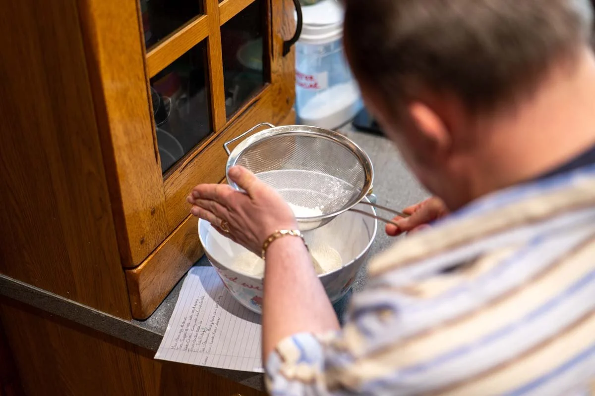 A man sifting flour into a bowl in a kitchen with wooden cabinets.