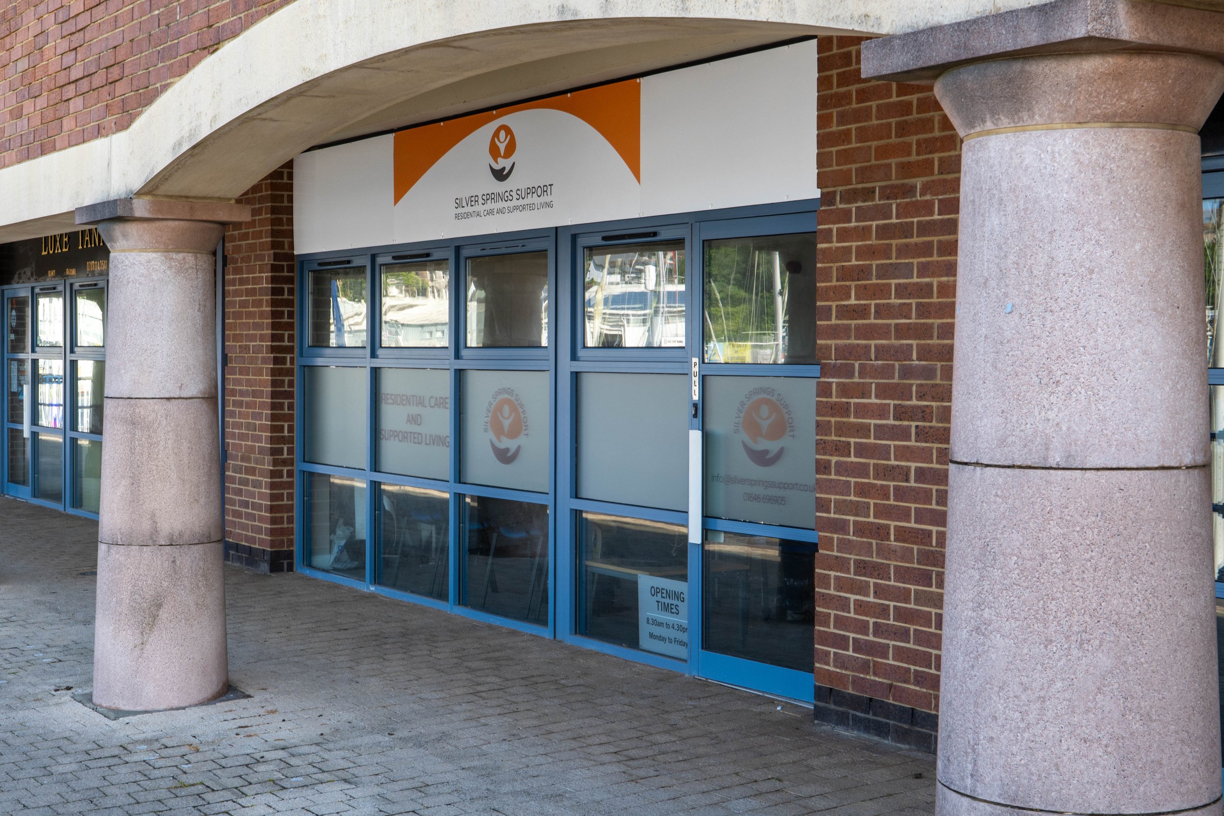 Storefront with brick walls and blue glass doors under an arched structure, supported by two large round stone columns; a sign reads 'Silver Springs Support' with details about residential care and supported living located in Milford Haven Waterfront