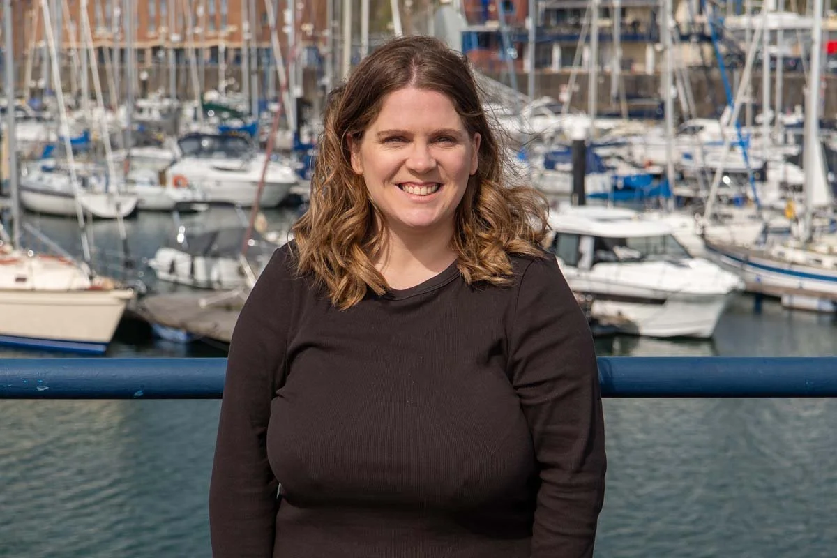 A woman with shoulder-length brown hair smiling in front of a marina with multiple sailboats docked.