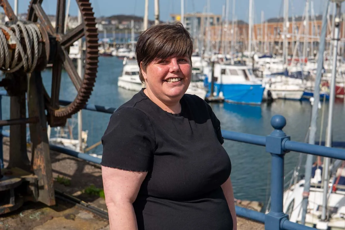 A woman with short brown hair smiling at the camera, standing near a marina with boats docked in the background.