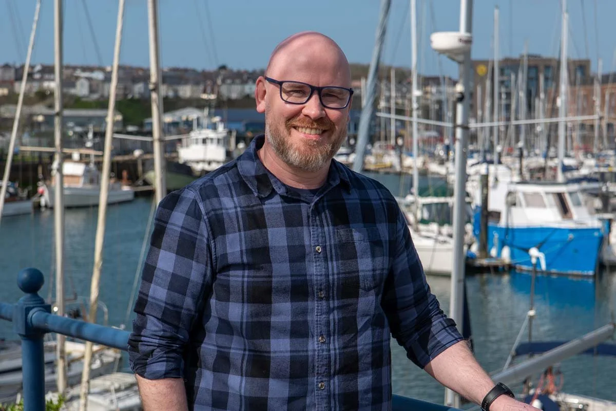 A smiling man with glasses and a beard in a blue plaid shirt standing near a marina with boats docked in the background.