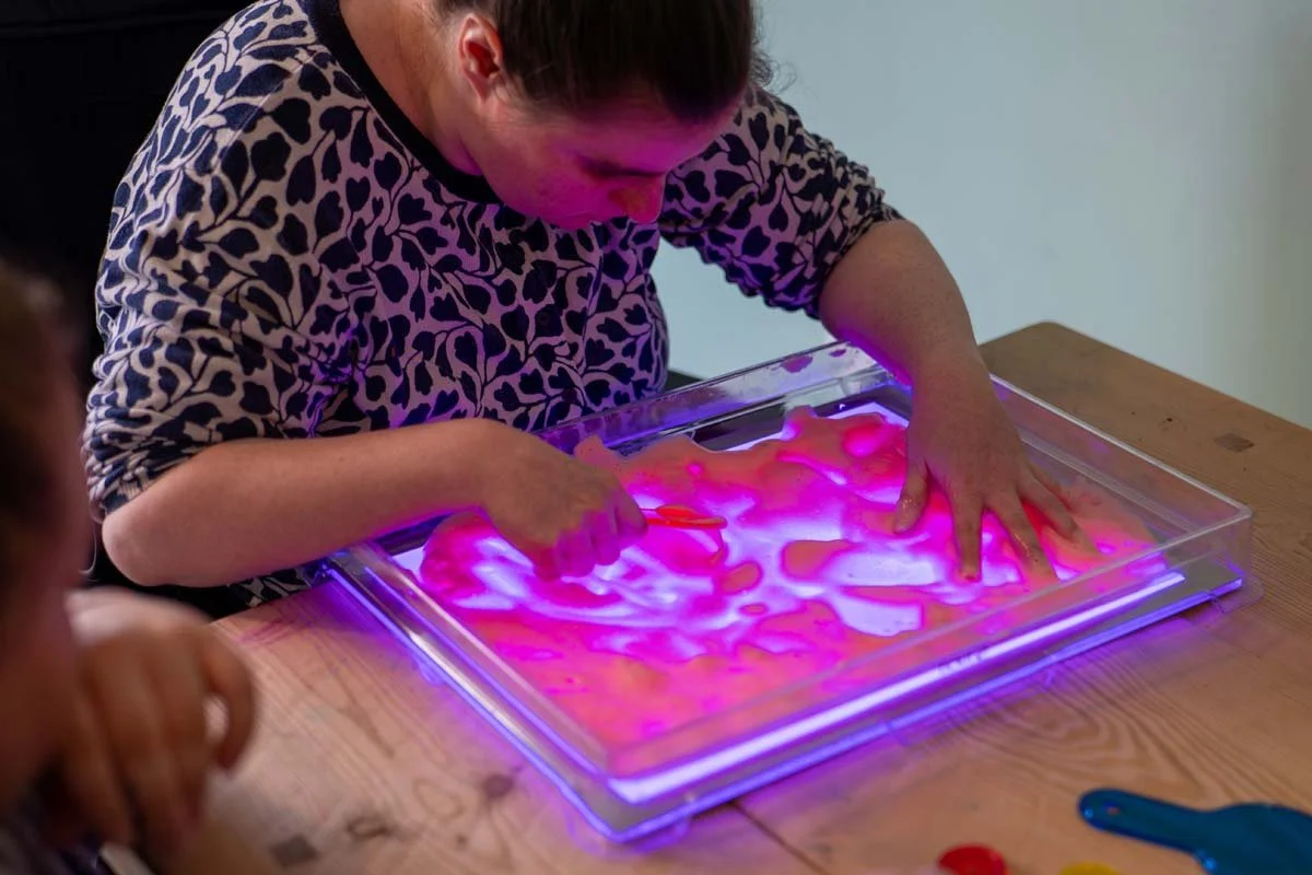 Child playing with a light-up sensory table, touching a surface illuminated with pink and purple colors.