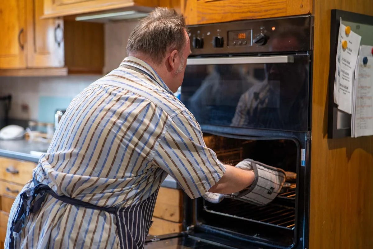 A man wearing a striped shirt and apron placing a dish into an oven in a kitchen.