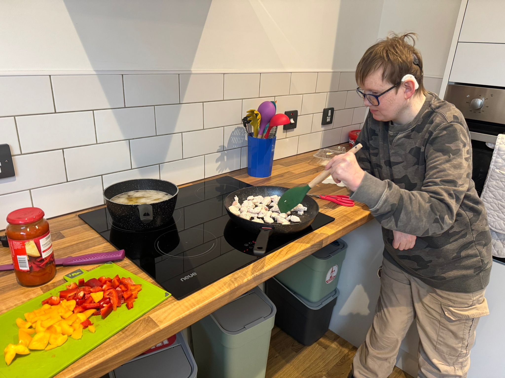 A young person with glasses and a disability hearing aid cooking chicken in a frying pan on a kitchen stove. The kitchen has white tiles, a counter with chopped red, yellow, and orange bell peppers, a jar of tomato sauce, and various colorful utensils in a blue container.