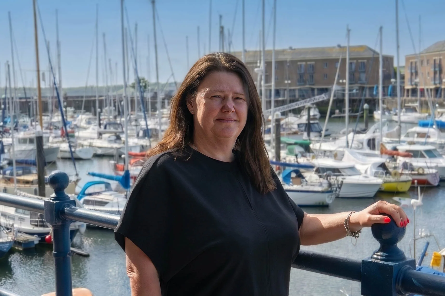 A woman with long brown hair, wearing a black shirt, standing on a marina dock with boats in the background and residential buildings.
