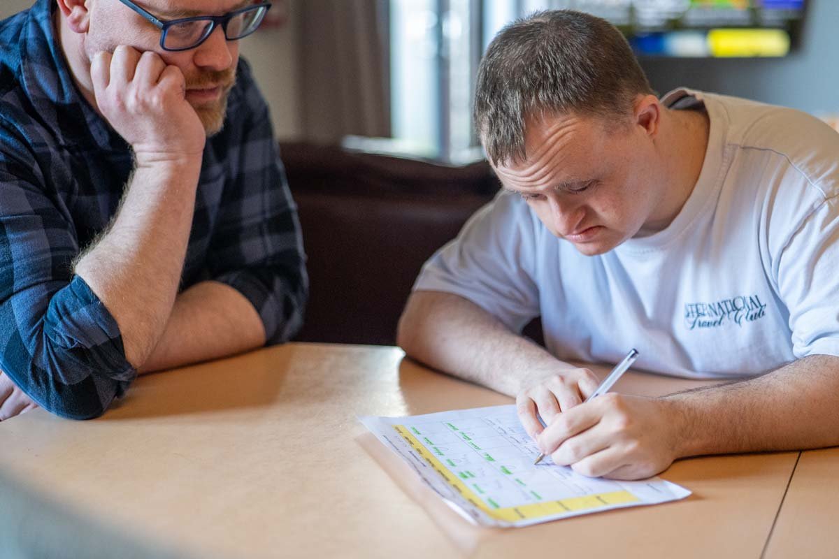 Two men seated at a wooden table, one writing and the other observing. The man on the left has glasses and a checked shirt, resting his head on his hand. The man on the right is in a white shirt and is writing on a sheet of paper with yellow highlighter and a pen in a well-lit indoor setting.