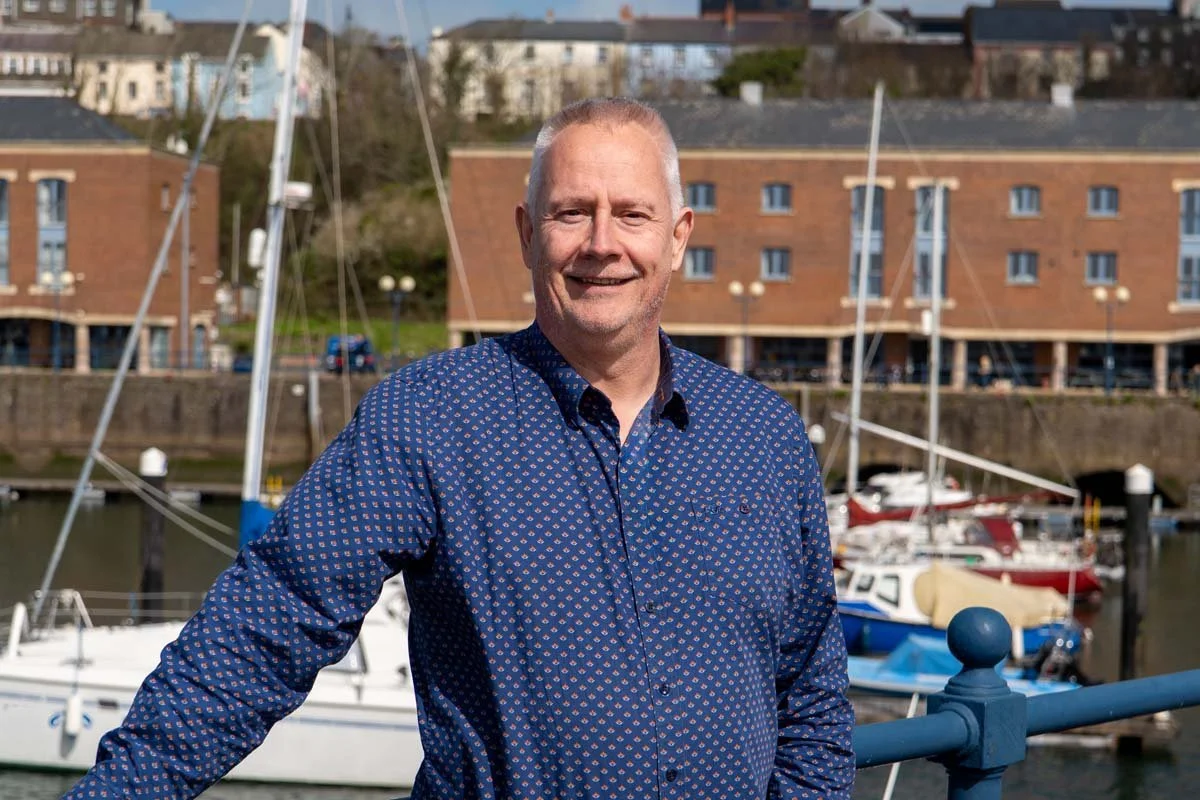 A smiling older man with short gray hair wearing a blue patterned dress shirt, standing near a marina with sailboats and a cityscape in the background.