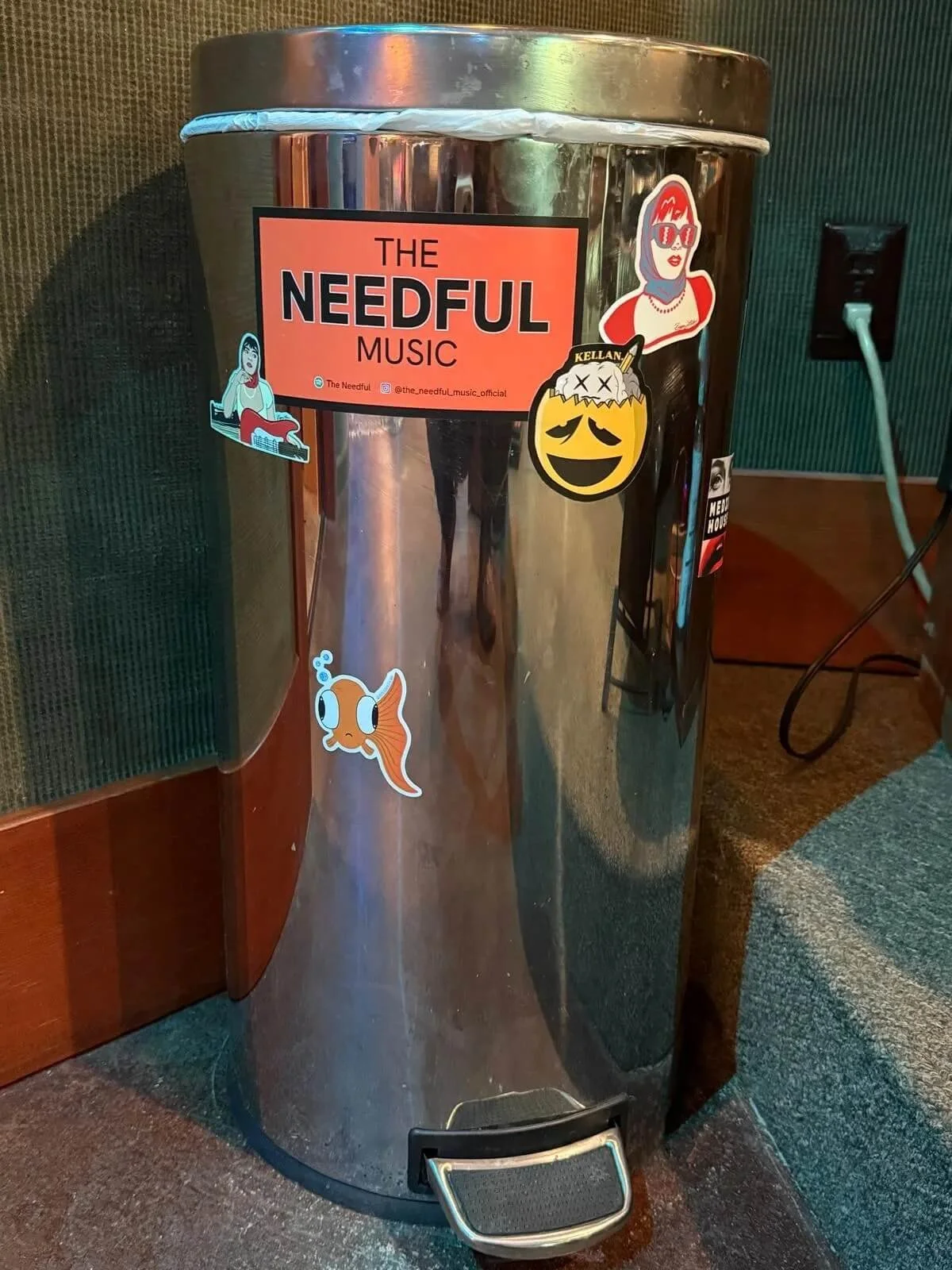 A metallic trash can with a label reading 'The Needful Music' and various stickers, including a cartoon woman with red hair, a smiling yellow face with sad eyes labeled 'Kellan', a cartoon fish, and a woman with sunglasses and red hair.