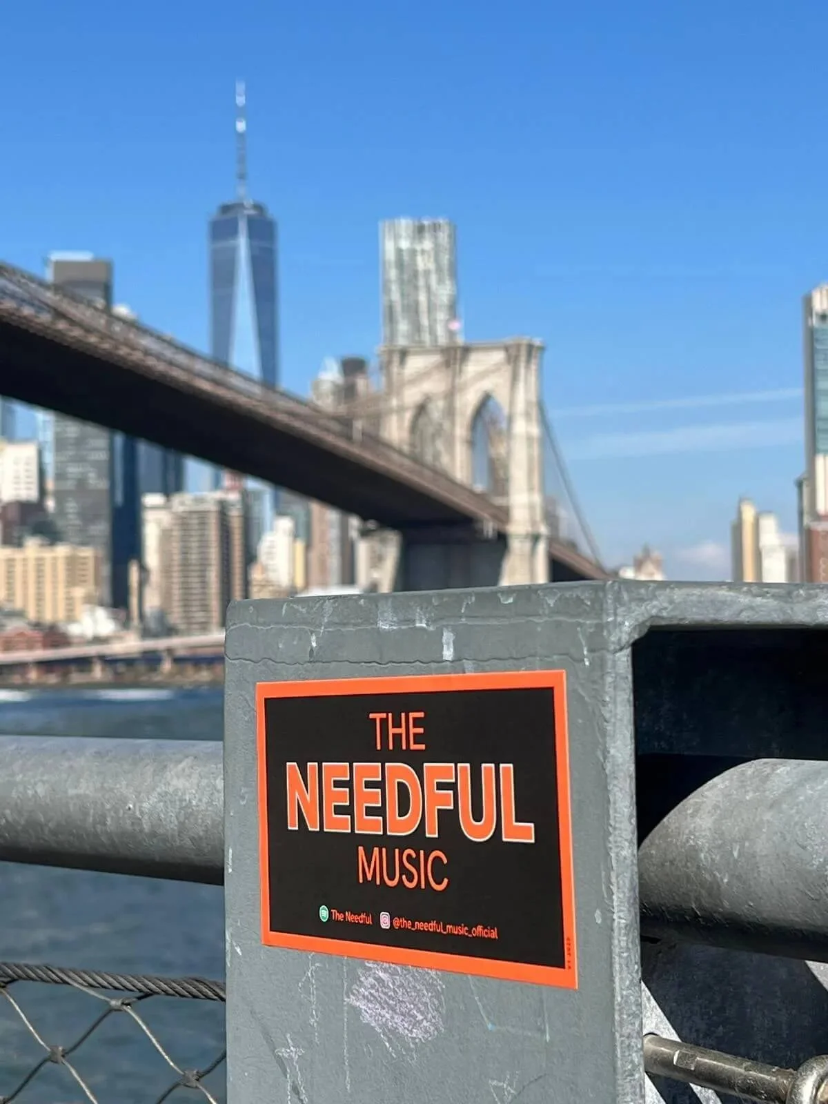 A sticker on a metal post that reads 'The Needful Music' with social media handles, set against the Brooklyn Bridge with the New York City skyline in the background on a clear, sunny day.