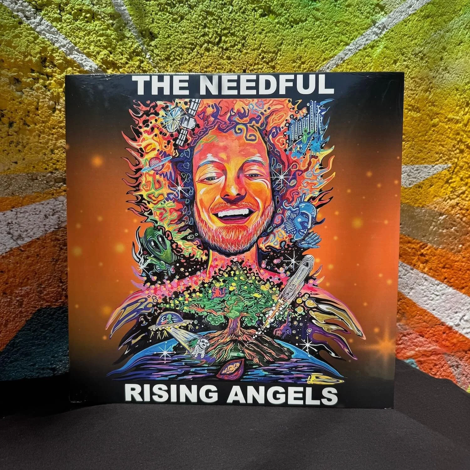 Colorful illustration of a smiling young man with a beard, surrounded by abstract, cosmic, and futuristic elements, with text 'THE NEEDFUL' at the top and 'RISING ANGELS' at the bottom.