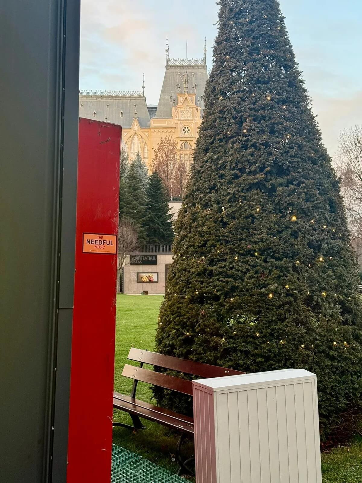 View of a Christmas tree with lights, a park bench, and a historic building with towers in the background, partially seen from behind a structure with a red panel in a city park.