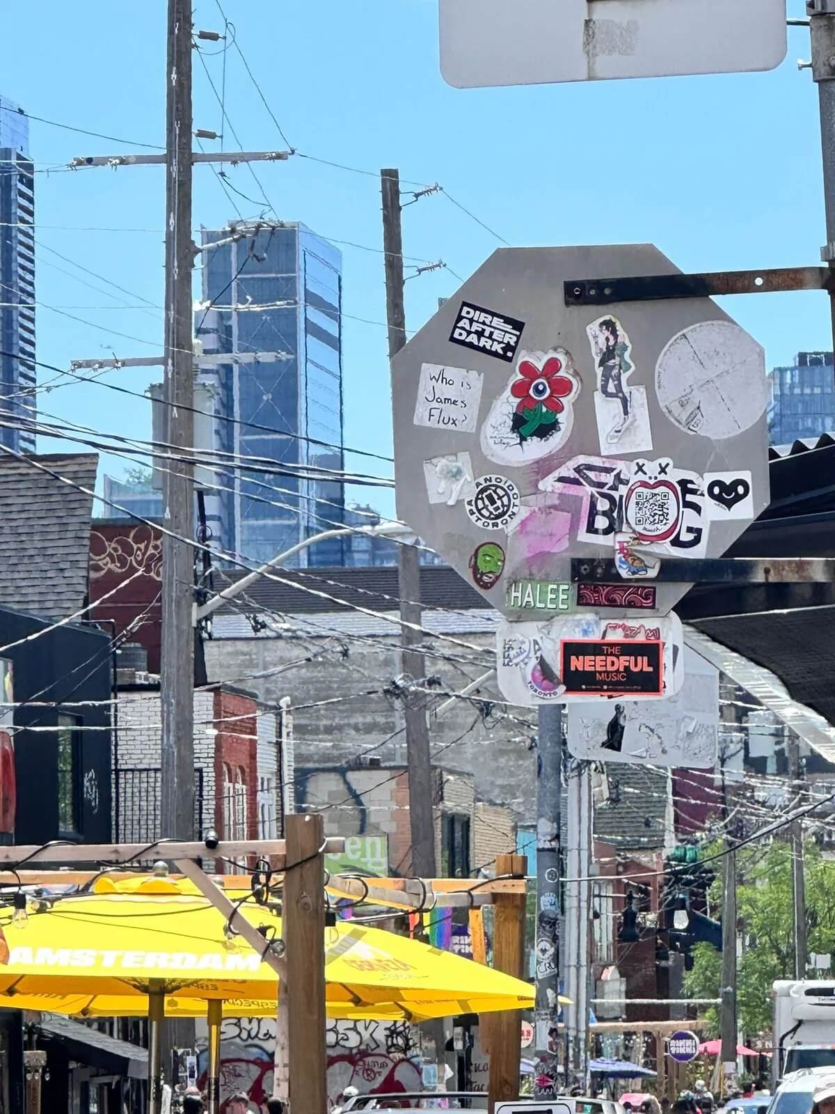 Street scene with utility poles, wires, and a collage of stickers on a street corner with buildings, umbrellas, and people in the background.