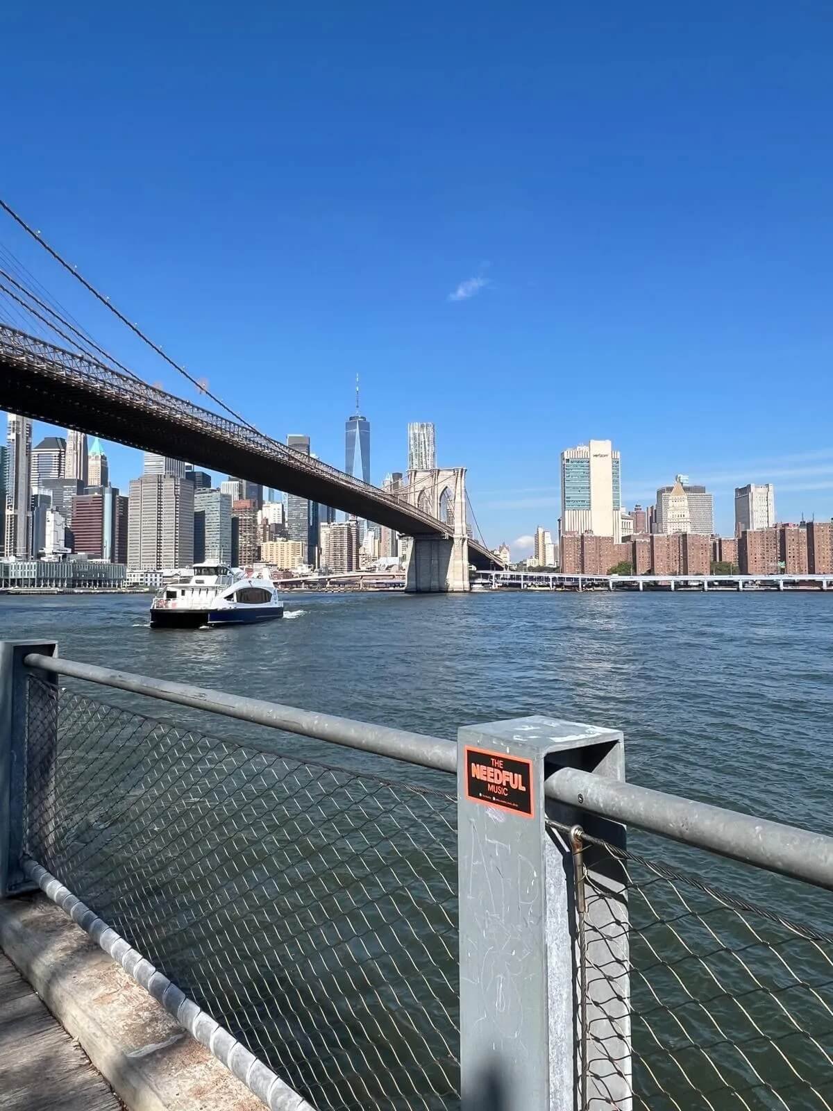 View of the New York City skyline with the Brooklyn Bridge crossing over the East River, and a boat sailing on the water in the foreground.