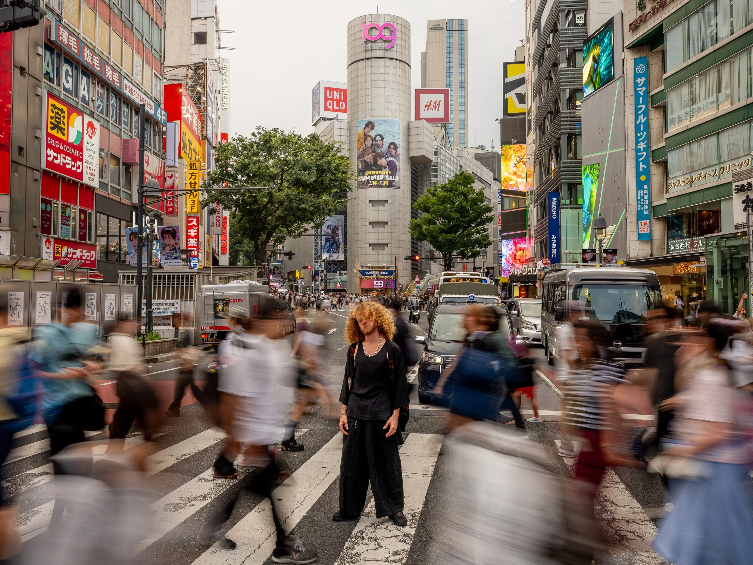 Busy city street scene in Tokyo with people crossing a crosswalk, tall buildings with colorful signs, billboards, and digital screens.