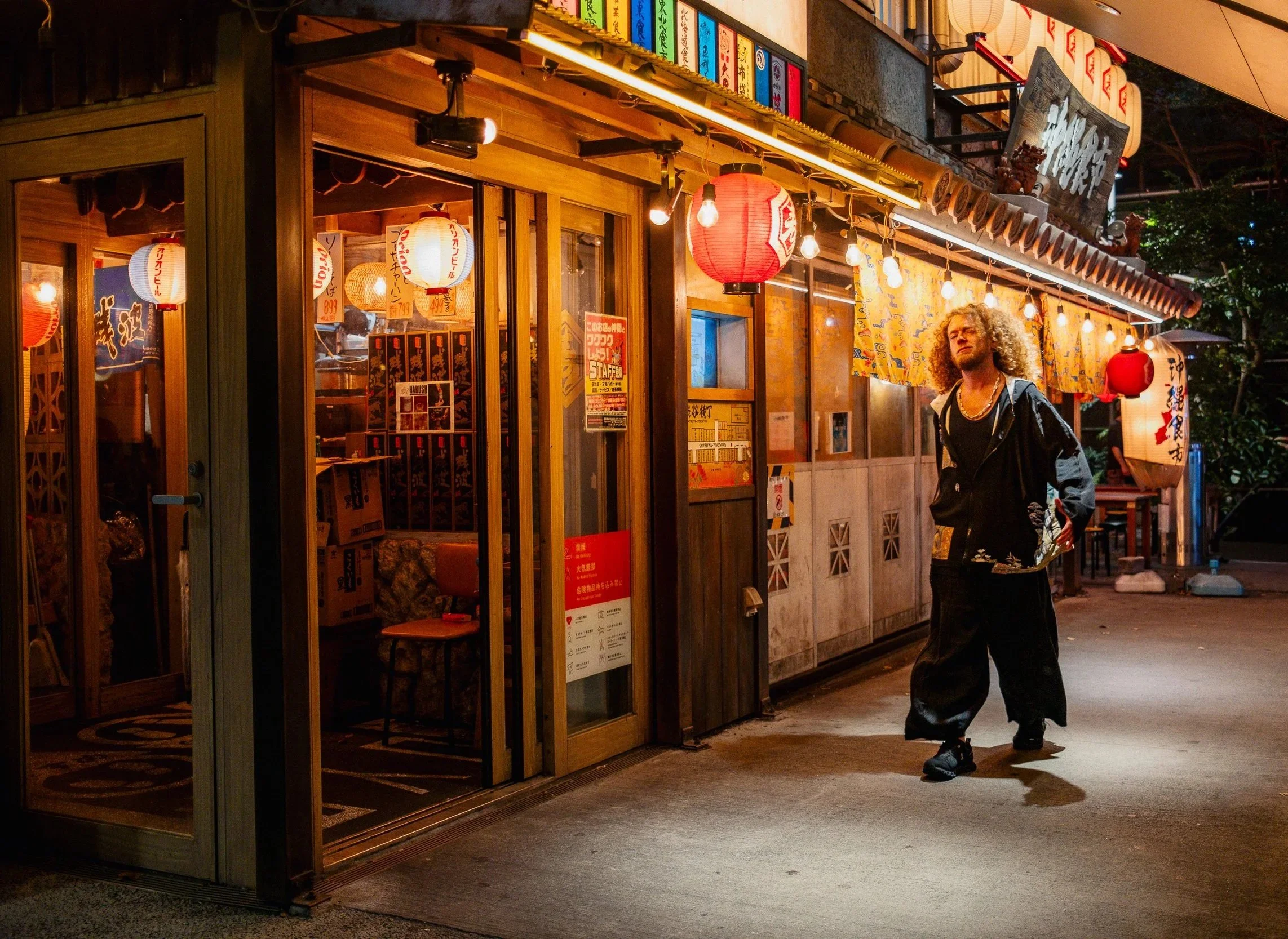 A man with curly hair wearing a black hoodie and baggy pants walking past a Japanese restaurant decorated with red lanterns and colourful signs at night.
