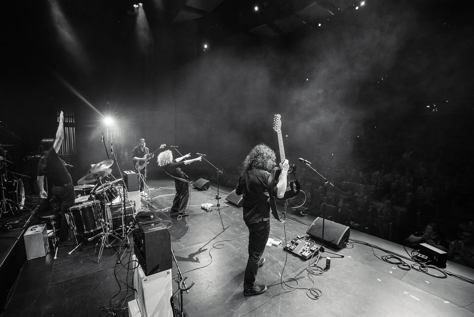 Black and white image of The Needful band performing on stage with various instruments, including drums, guitars, and microphones, under stage lighting, and an audience visible in the background.