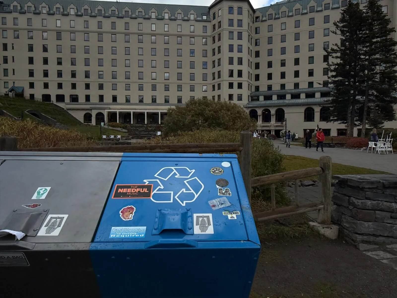 Blue recycling bin with stickers, outdoor scene with people walking near a large white building, trees, and benches.