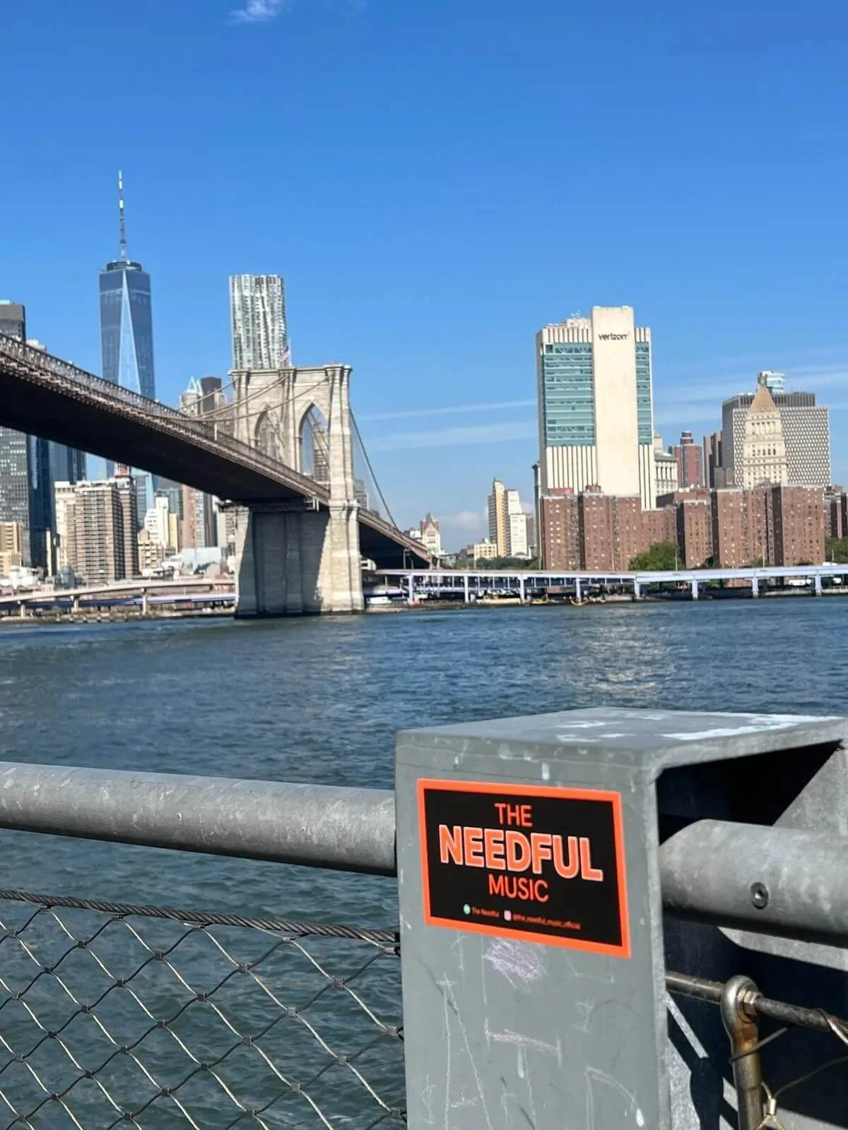 City skyline featuring the Brooklyn Bridge and various skyscrapers, with a body of water in the foreground, and a metal railing with a sticker reading 'The Needful Music'.
