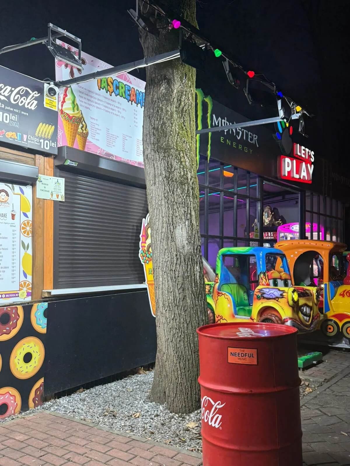 Colorful amusement park with kiddie rides, a crate with a label that reads 'The Needful Music,' a Coca-Cola barrel, a tree, and an ice cream stand at night.