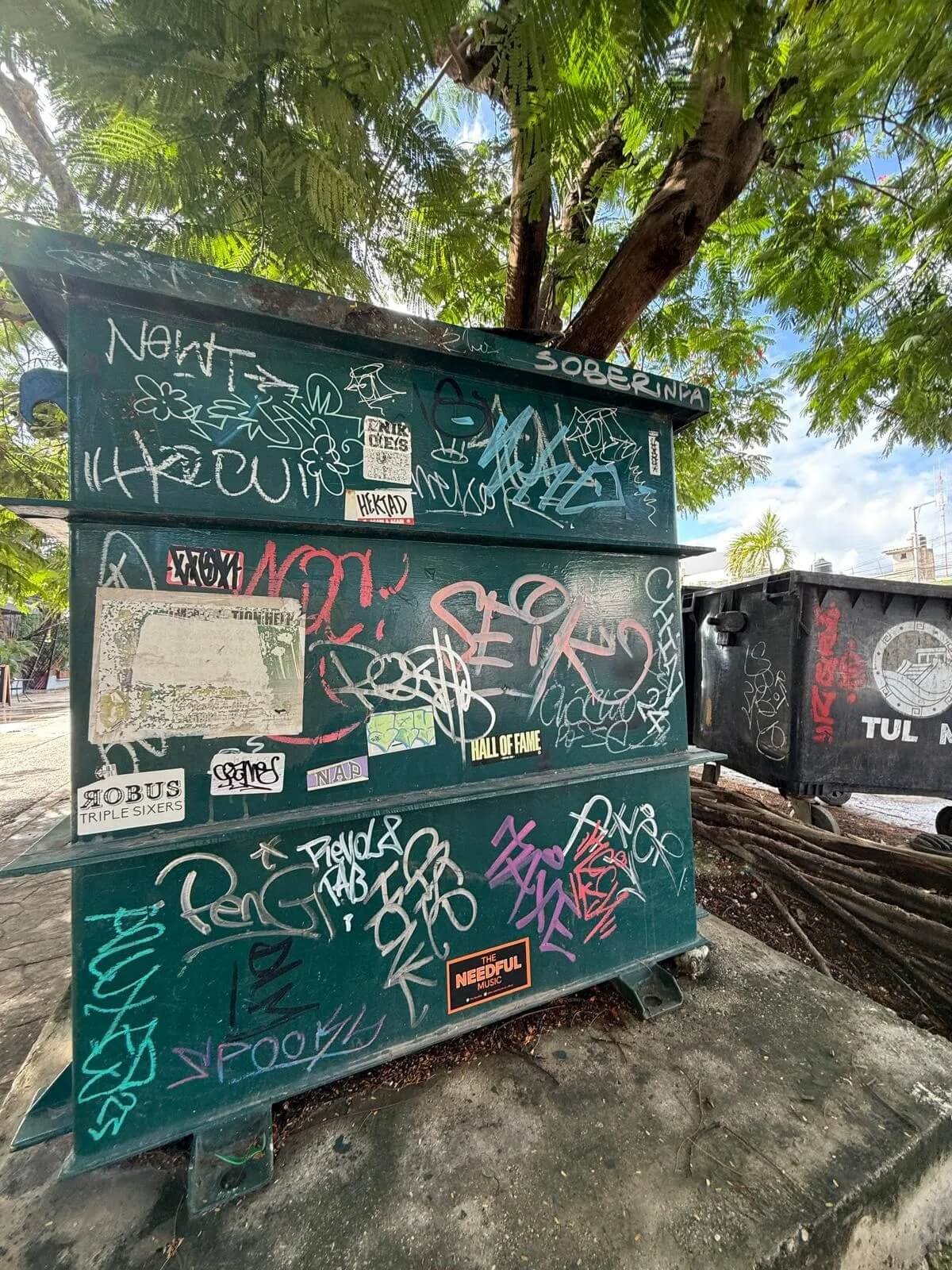 Green electrical box covered in various colorful graffiti and stickers, positioned outdoors under a tree.
