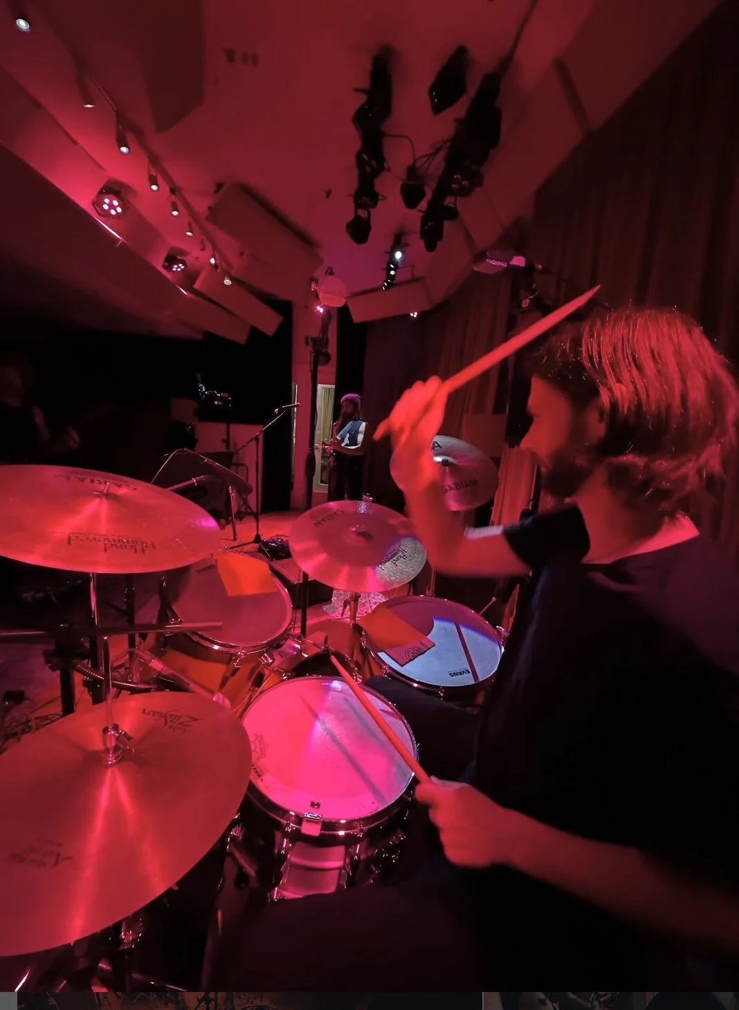 Stefan of The Needful playing drums during a performance in a dimly lit venue with red lighting, a drummer holding drumsticks, and a musician in the background.