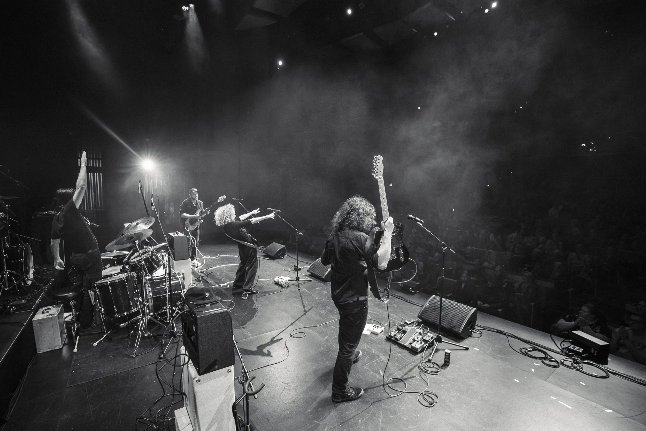 Black and white photo of a live musical performance by The Needful on stage with Trooper, with multiple musicians playing instruments including drums and guitar, with stage lights illuminating the scene.